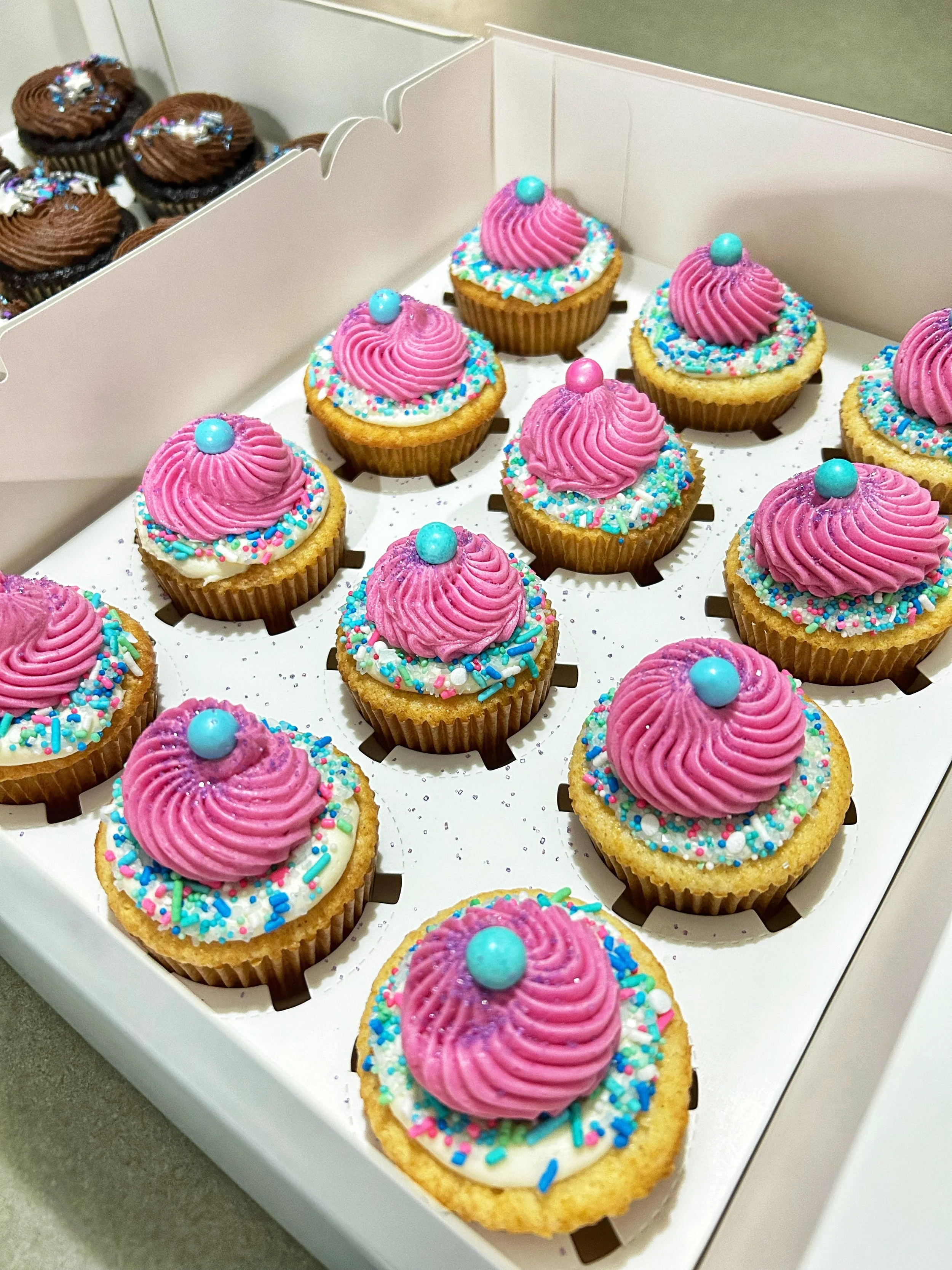 A tray of decorated cupcakes with pink frosting, colorful sprinkles, and a blue candy on top, with chocolate cupcakes in the background.