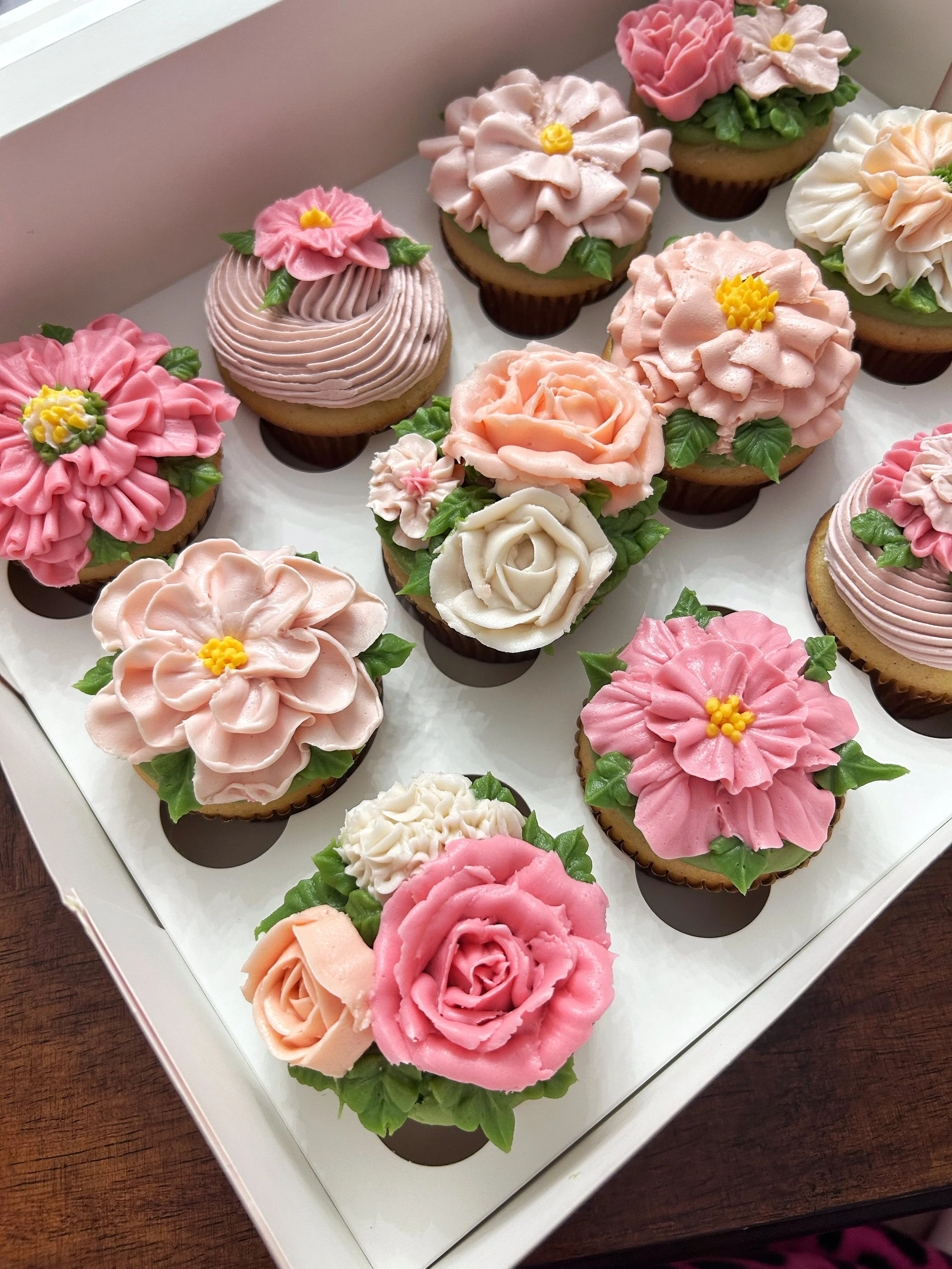 A box of assorted cupcakes decorated with floral themes in pink, white, and peach colors with green leaves.