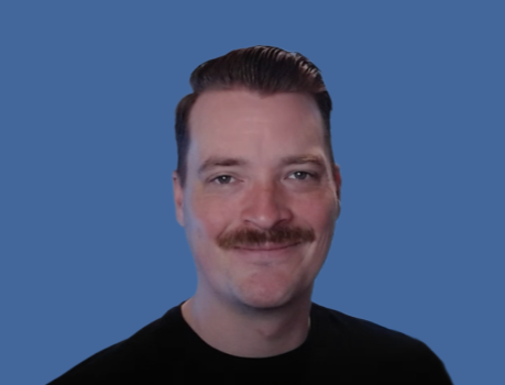 A man with a mustache and slicked-back hair smiling at the camera against a blue background.