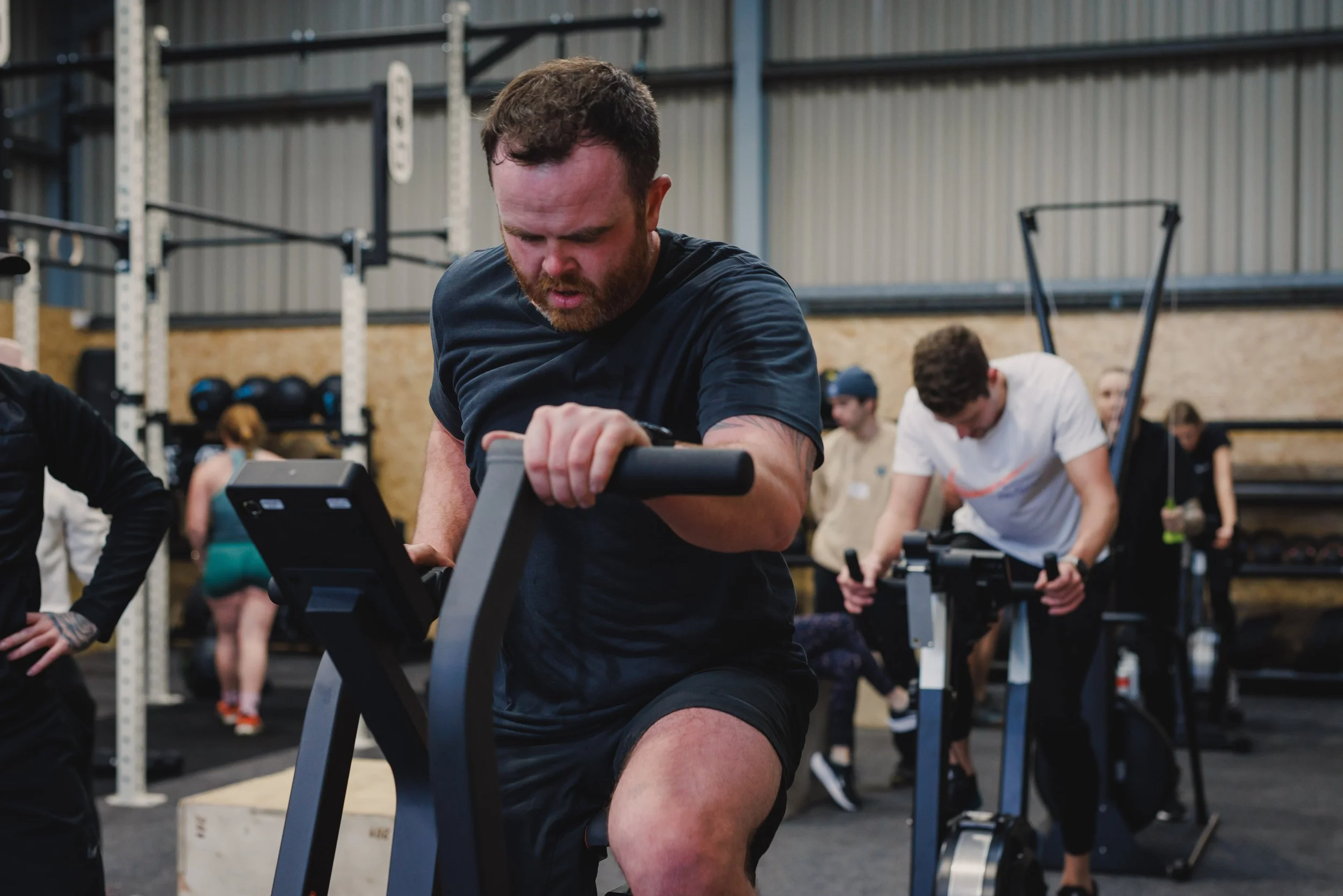Man working out on an echo bike at a gym, with other people exercising in the background.