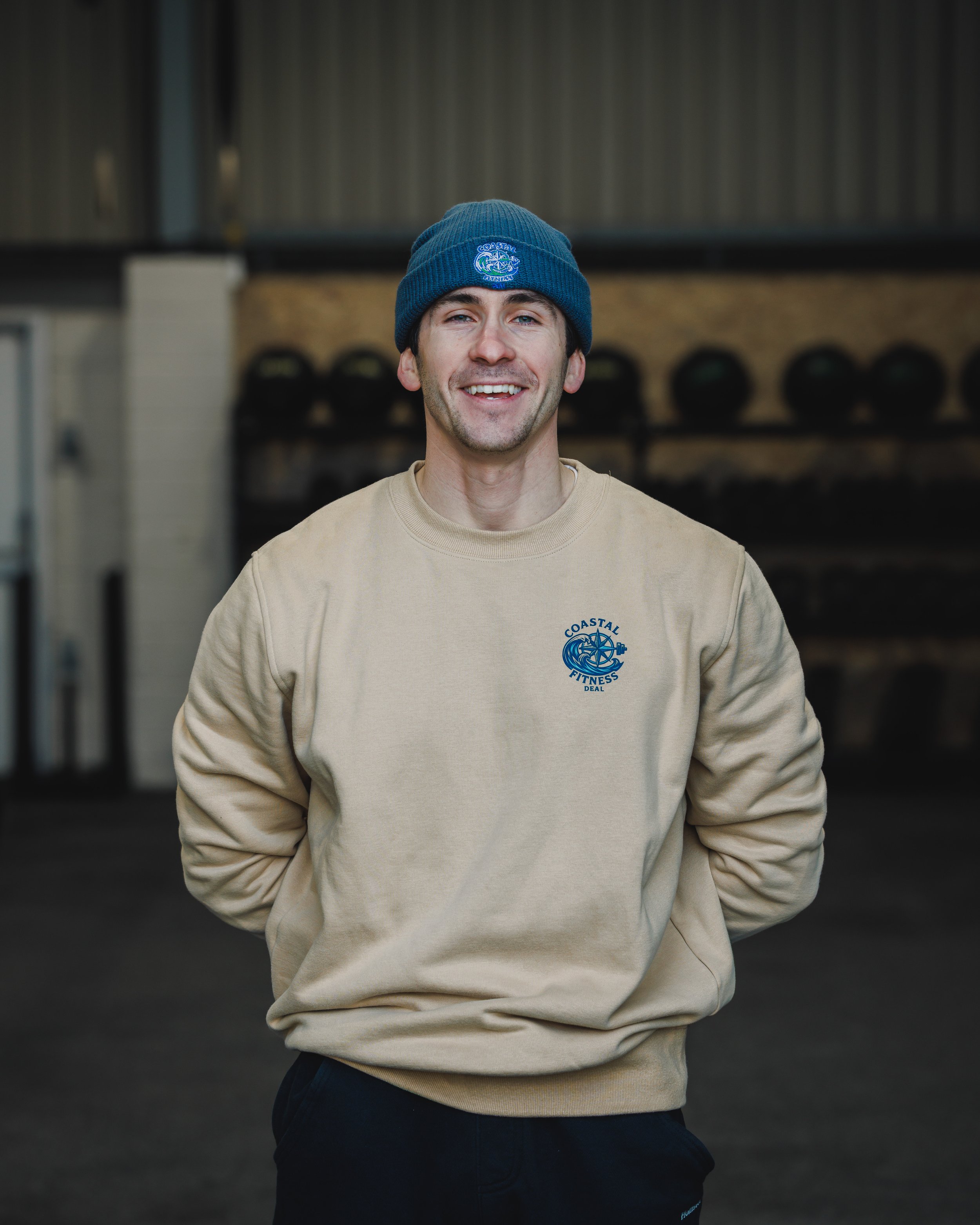 A man smiling, wearing a beige sweatshirt with a blue logo that reads 'Coastal Fitness Deal' and a blue beanie, standing in an indoor gym.