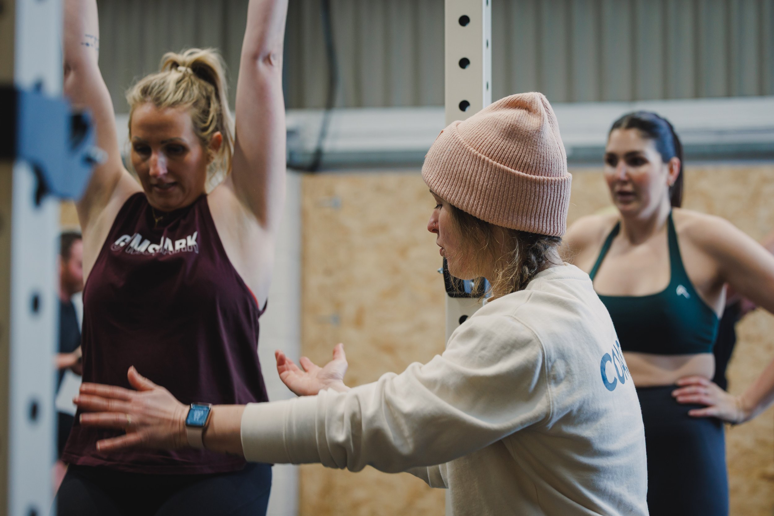 A woman in a maroon tank top is lifting weights while a trainer in a beige beanie and sweatshirt guides her on form. Two other women in athletic wear are visible in the background.