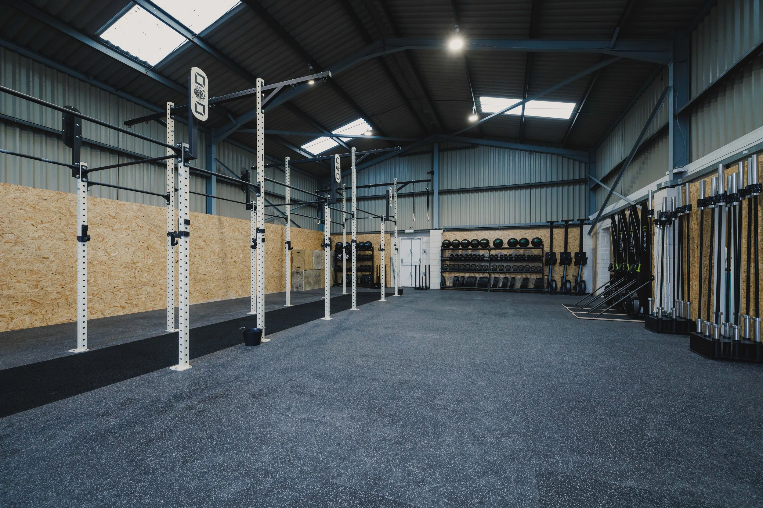 Empty gym with white weight racks, black and green weights, black rubber flooring, and a high ceiling with skylights.