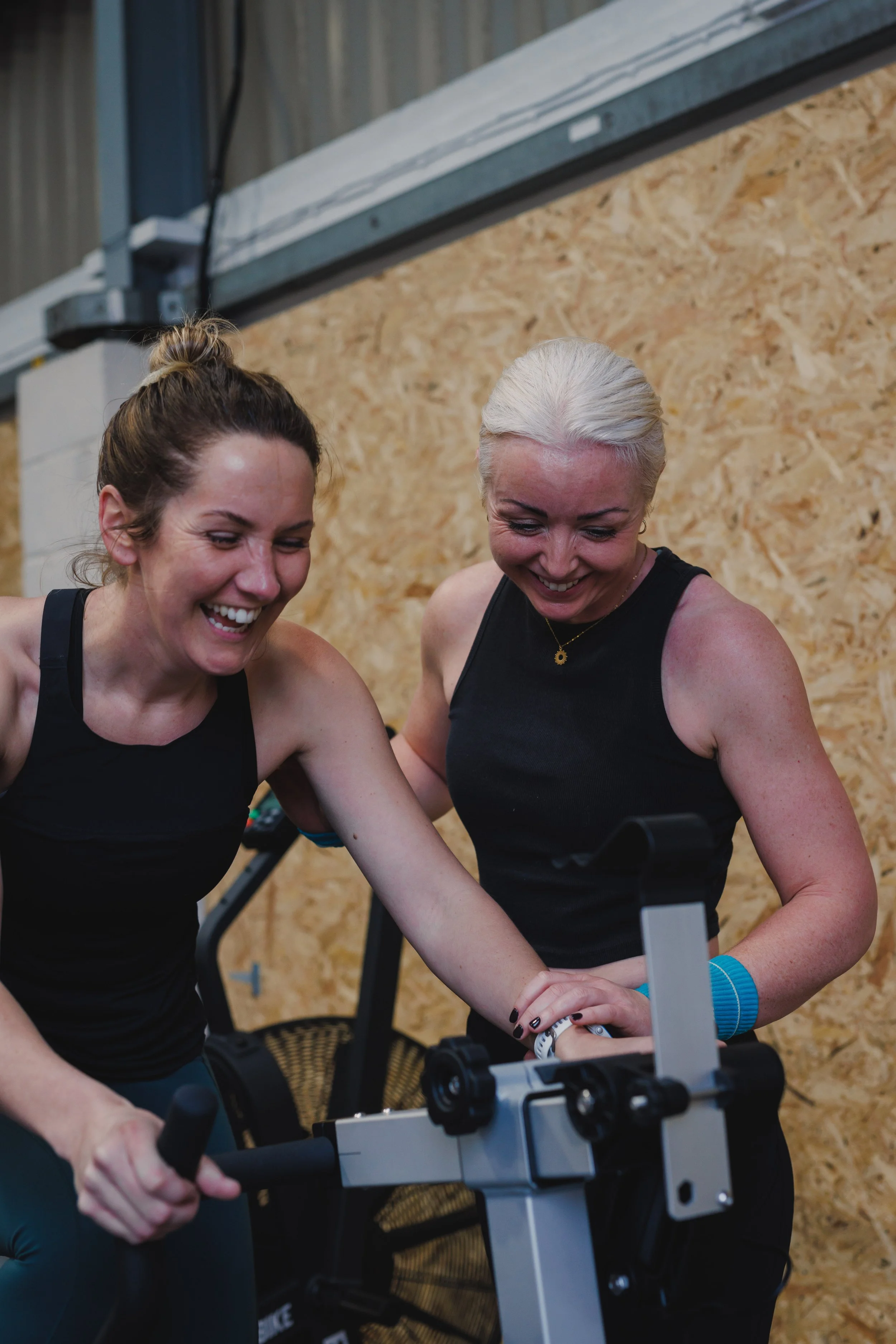 Two women smiling and laughing during a workout session at the gym, one on a stationary bike, with a wooden wall in the background.