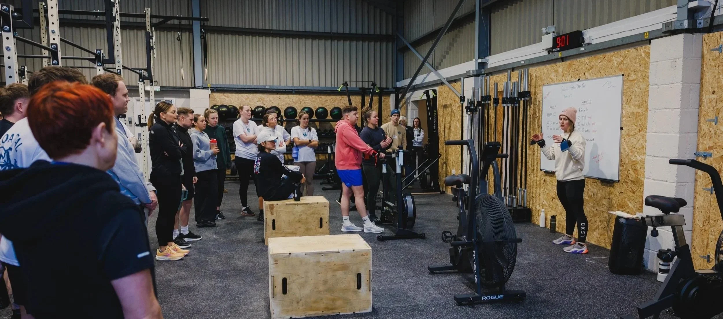 A group of women and girls listening to a woman trainer in a gym. The trainer is standing in front of a whiteboard and explaining. Gym equipment and black medicine balls are visible in the background.