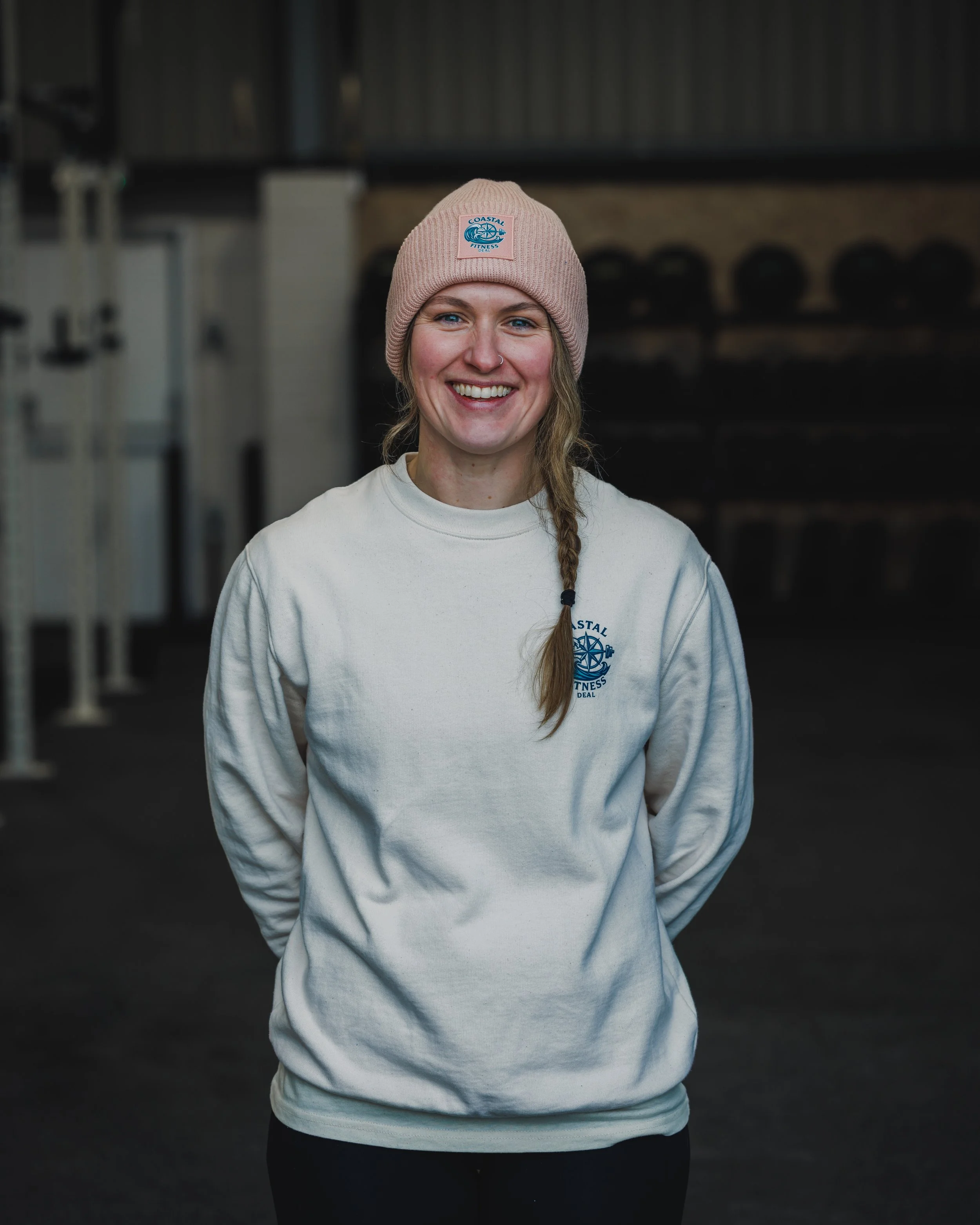 A smiling young woman with a braid, wearing a pink beanie and a white sweatshirt, standing inside a gym.