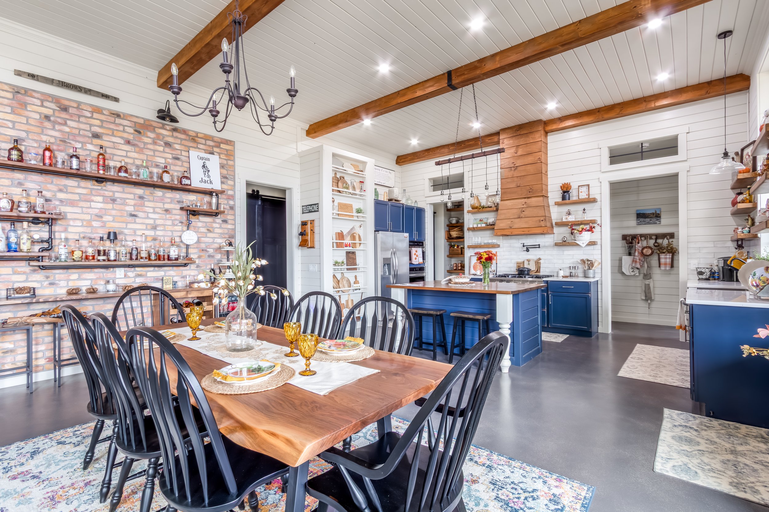 A spacious kitchen and dining area with a rustic and modern decor. It features a large wooden dining table with black chairs, a brick accent wall with shelves and liquor bottles, and a blue kitchen island with stools. The white walls and ceiling have
