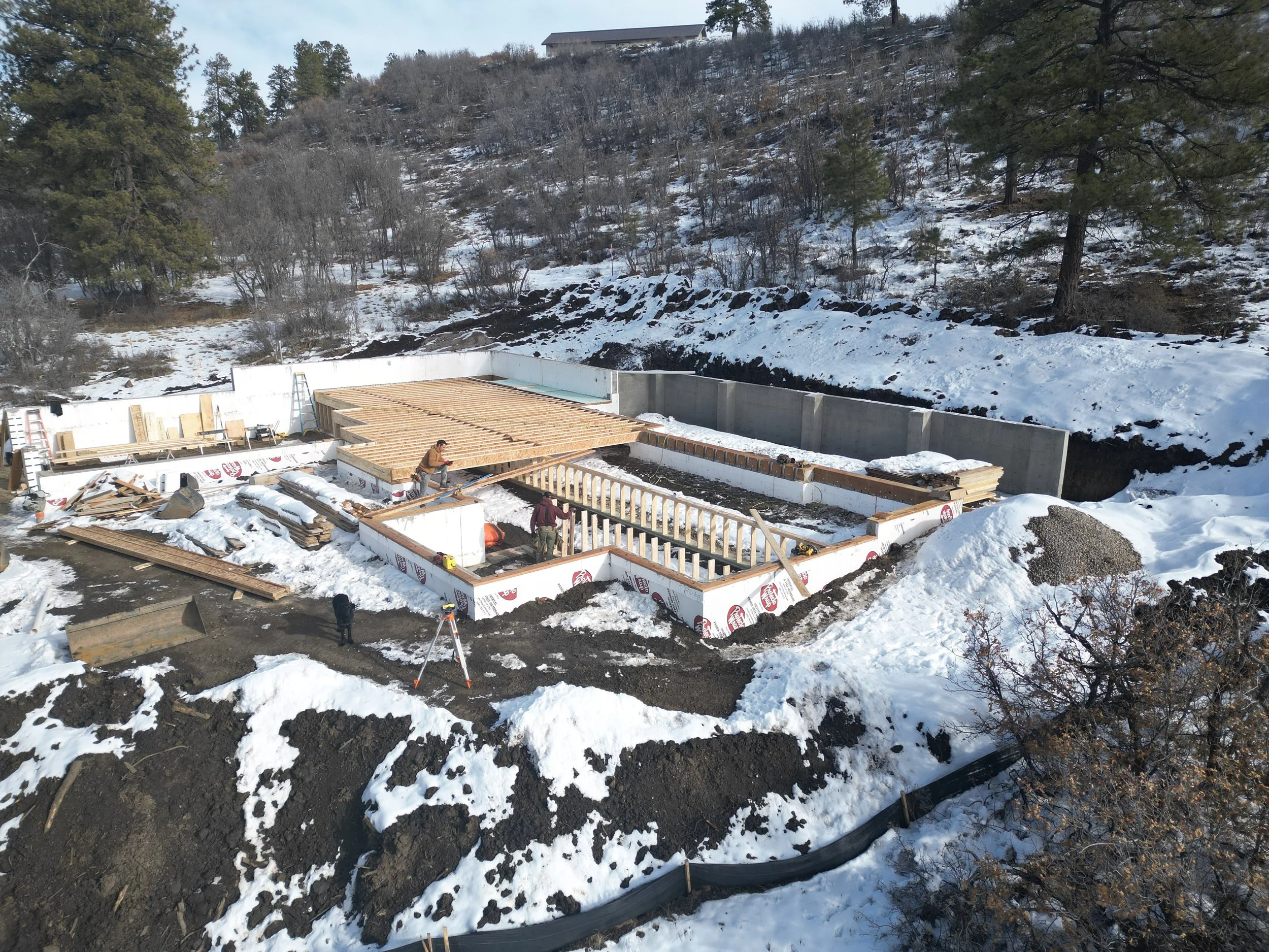 Construction site in a snowy landscape with workers building a house, wooden framework, and exposed foundation walls, surrounded by trees and mountains.
