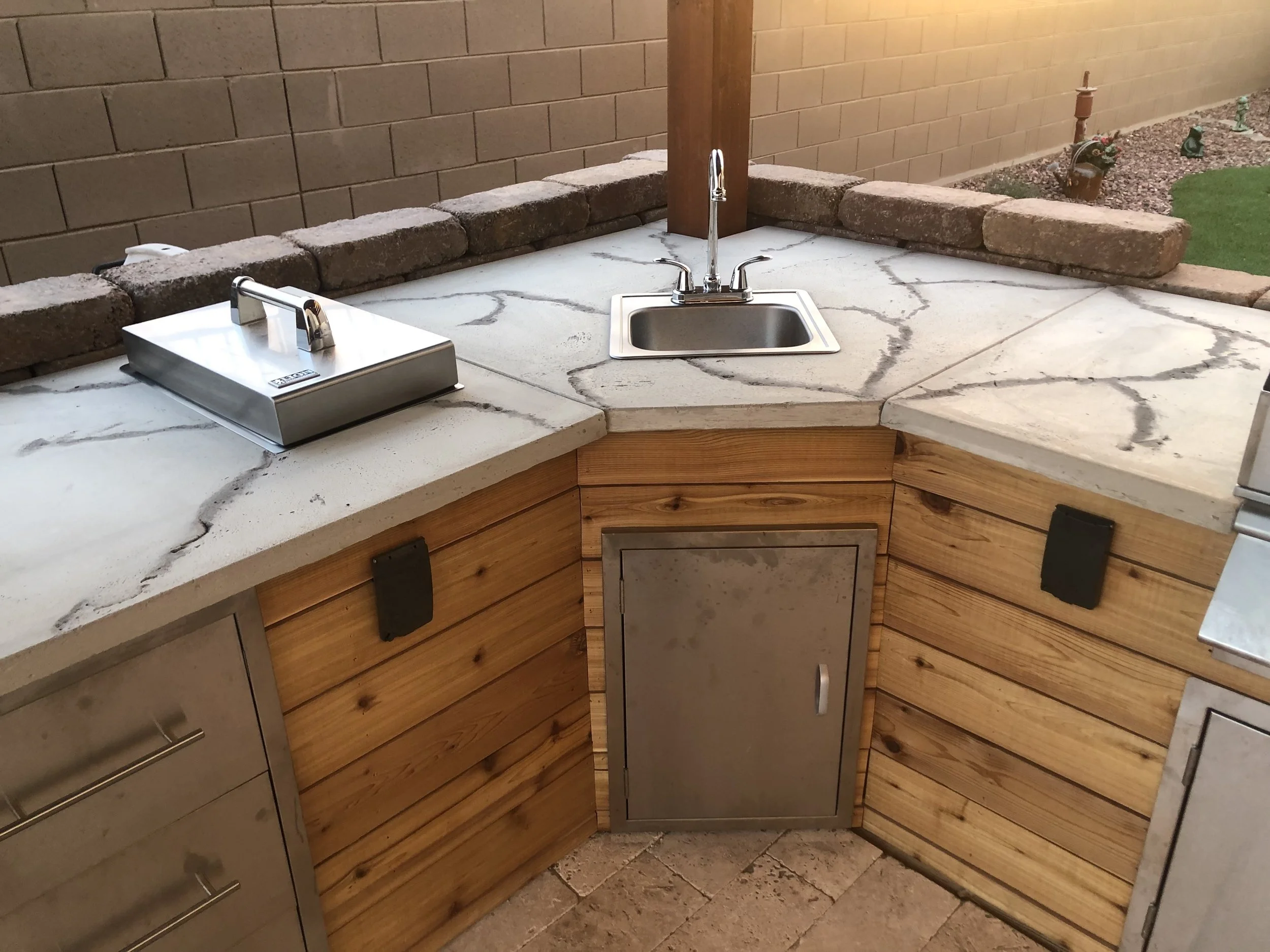 Outdoor kitchen with a stainless steel sink, a hot dog roller grill, and a marble countertop, surrounded by brick and wood paneling.
