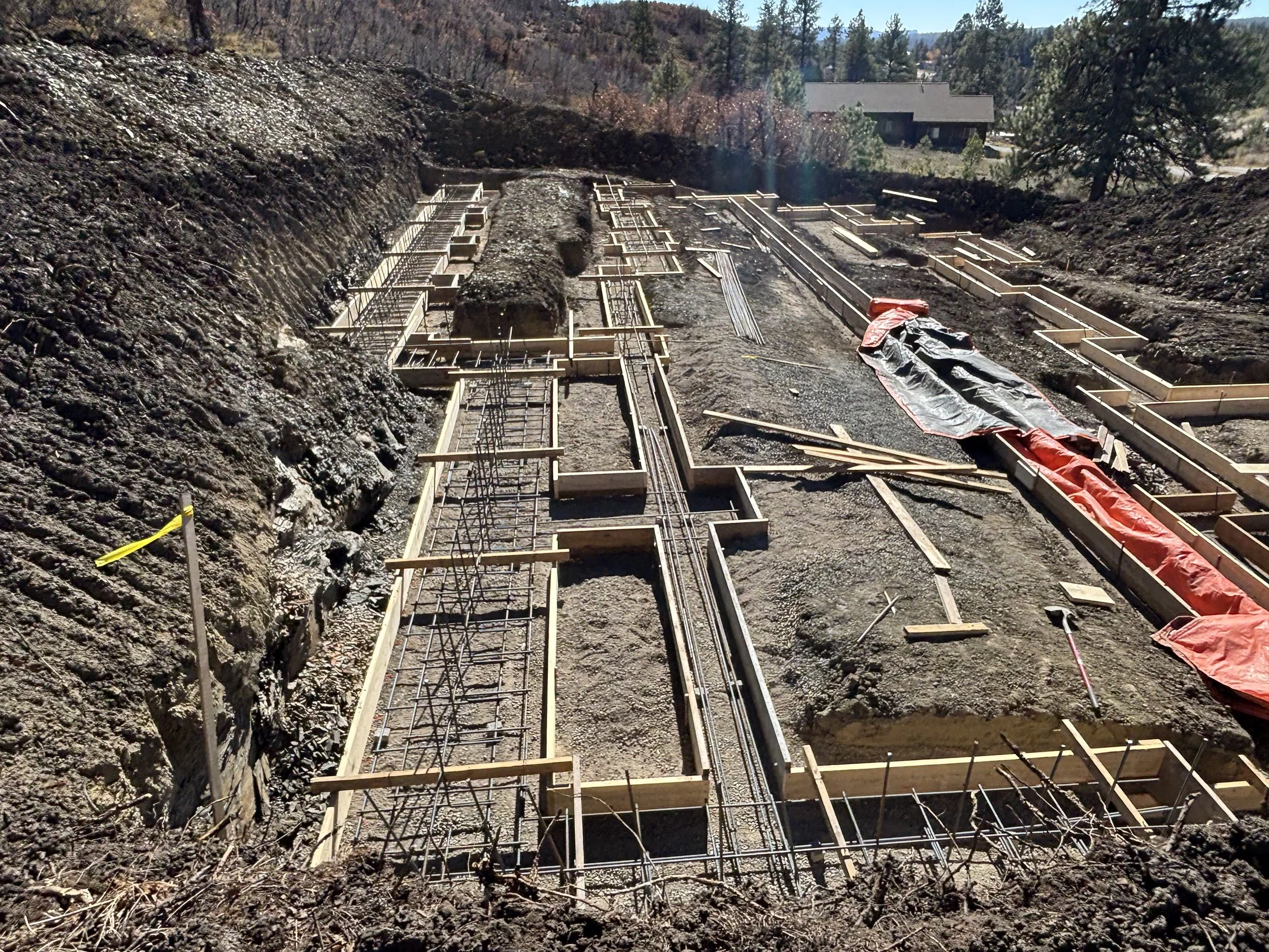 Construction site with wooden formwork and steel rebar for building foundation, working in an outdoor area with dirt and soil, residential houses and trees visible in the background.