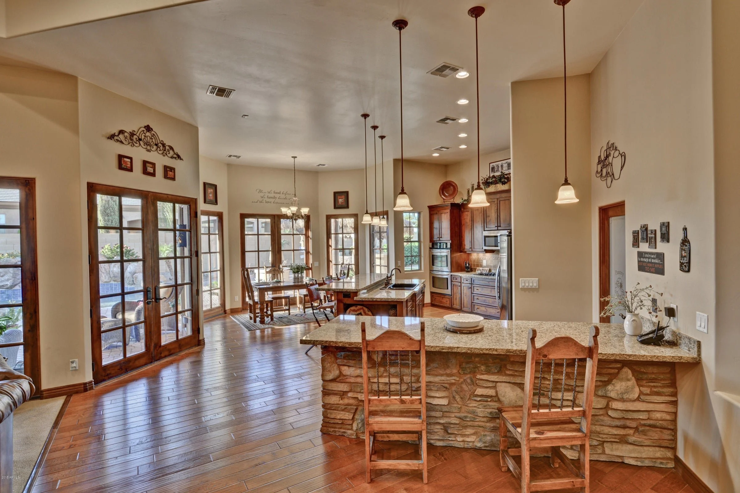 A spacious kitchen and dining area with wooden floors, granite countertops, wooden cabinets, and large windows allowing natural light; includes a kitchen island, two wooden chairs, and a dining table with chairs.