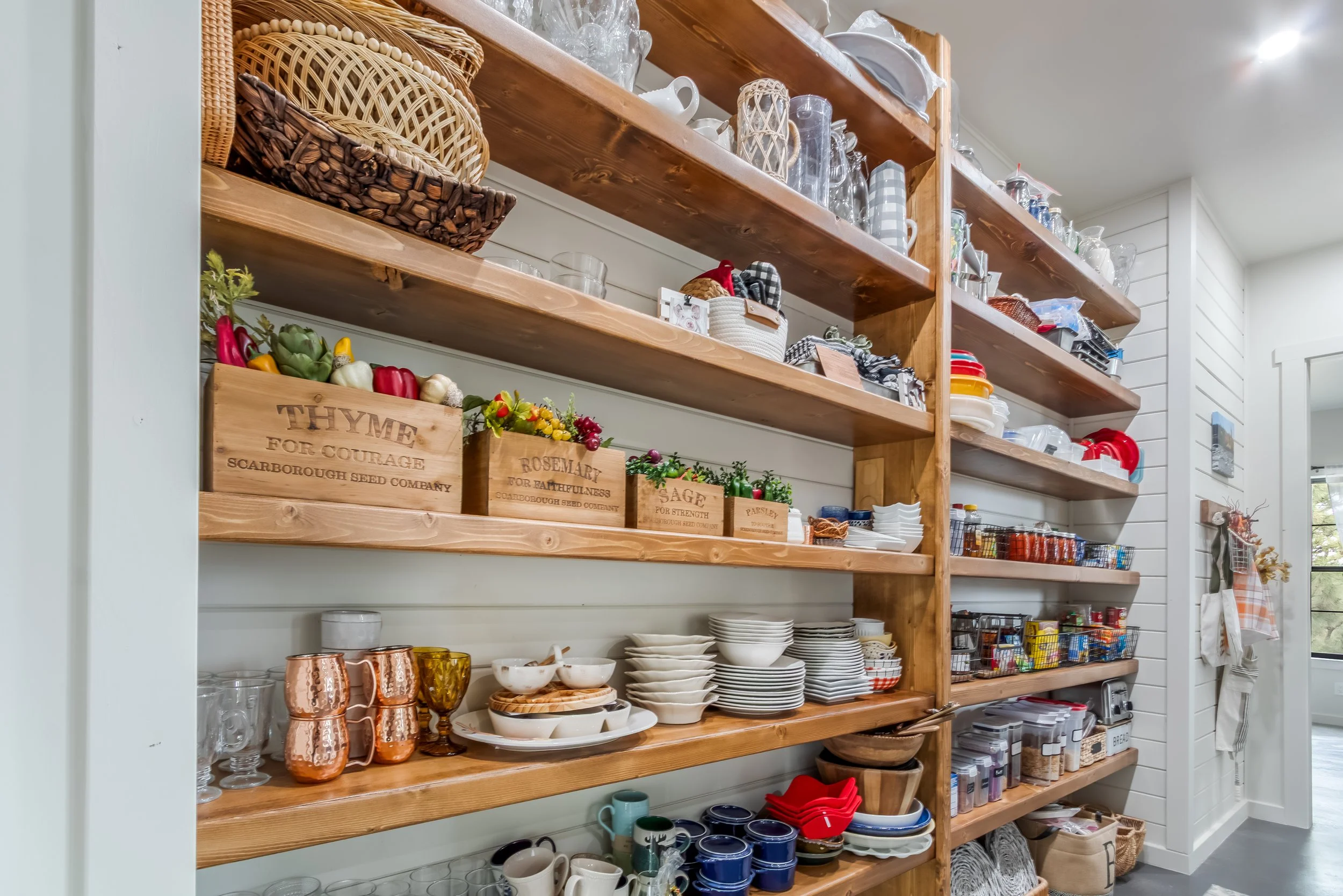 Open wooden shelves in a kitchen filled with plates, bowls, glasses, cups, and kitchen accessories, with some decorative crates and small plants.