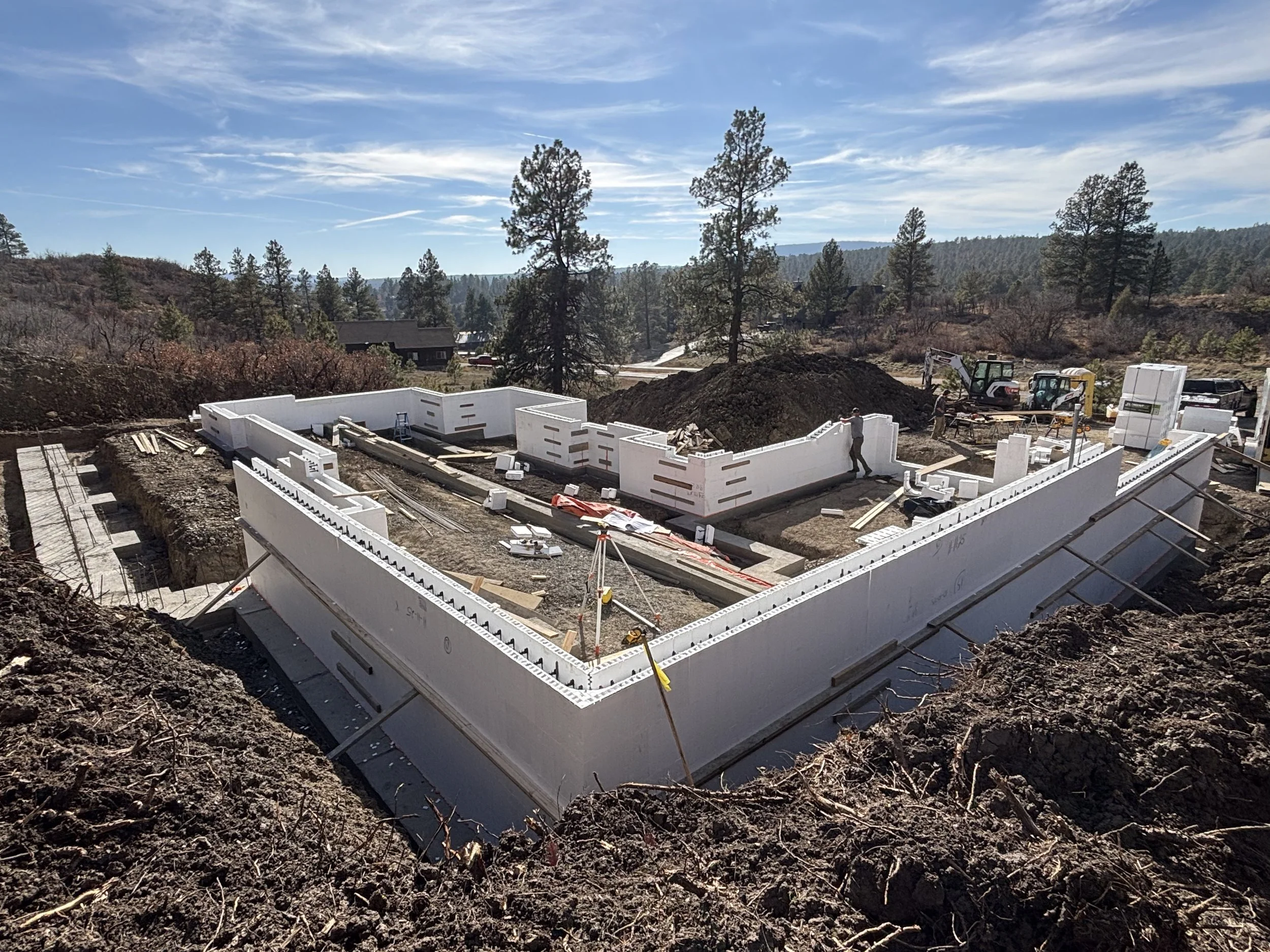 Construction site with insulated wall panels being assembled for a building foundation, surrounded by dirt and construction equipment, set against a backdrop of hills and pine trees under a partly cloudy sky.