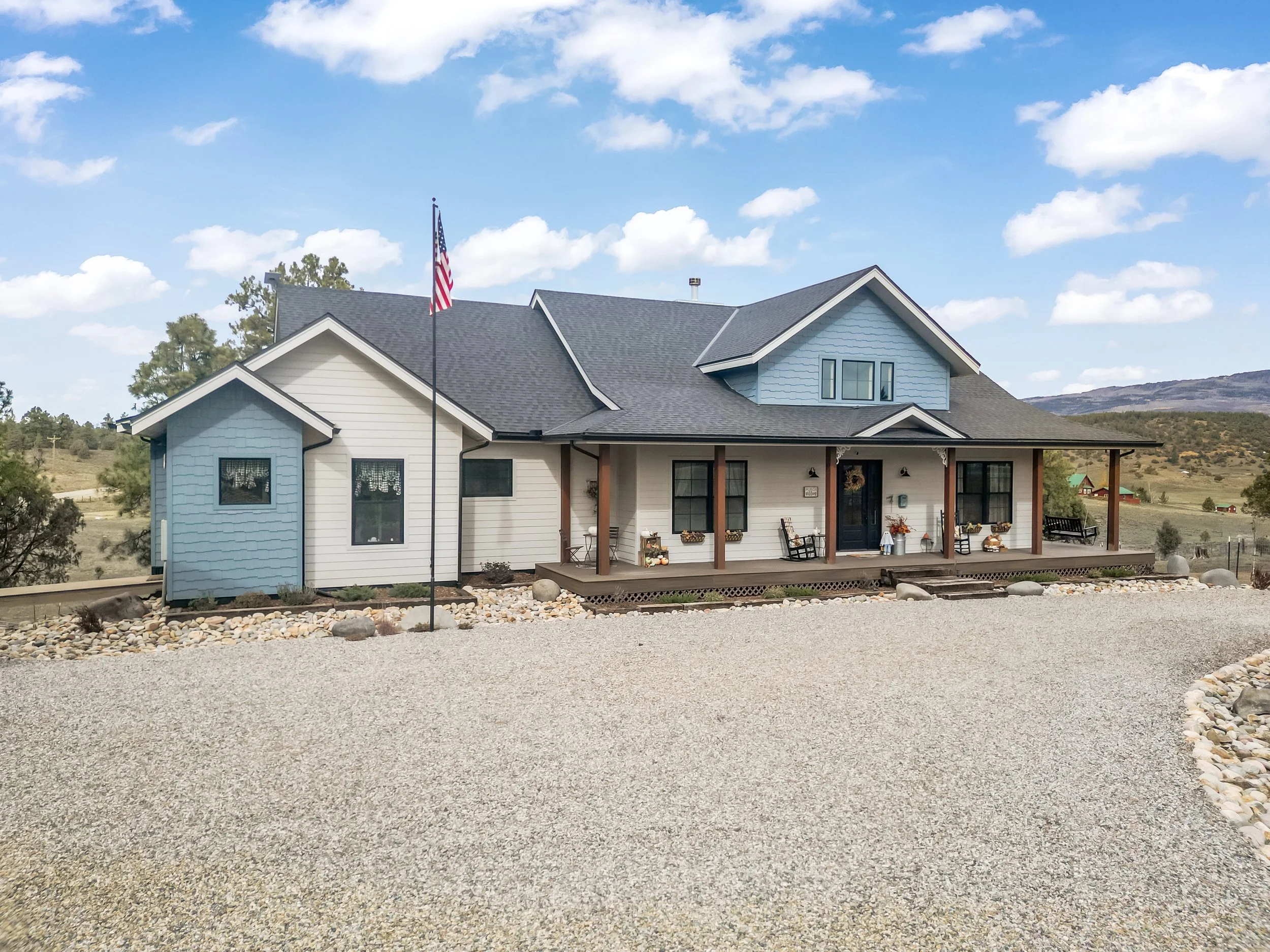 A modern house with a mix of white and light blue exterior siding, black window frames, and a dark gray roof, surrounded by a gravel driveway under a partly cloudy sky.