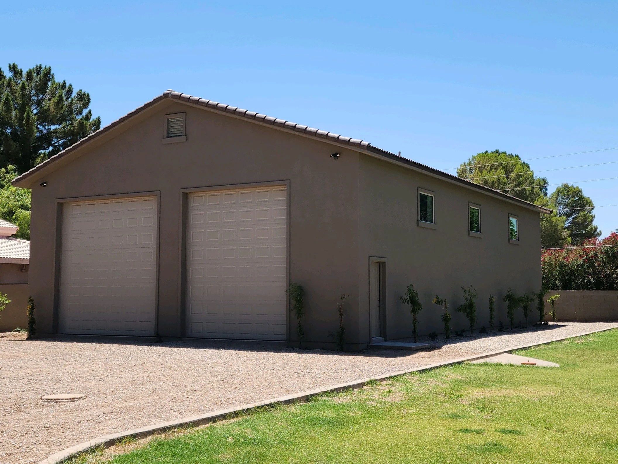 A beige two-story garage with two large garage doors and a side door, surrounded by a gravel driveway and green grass, with trees and a clear blue sky in the background.