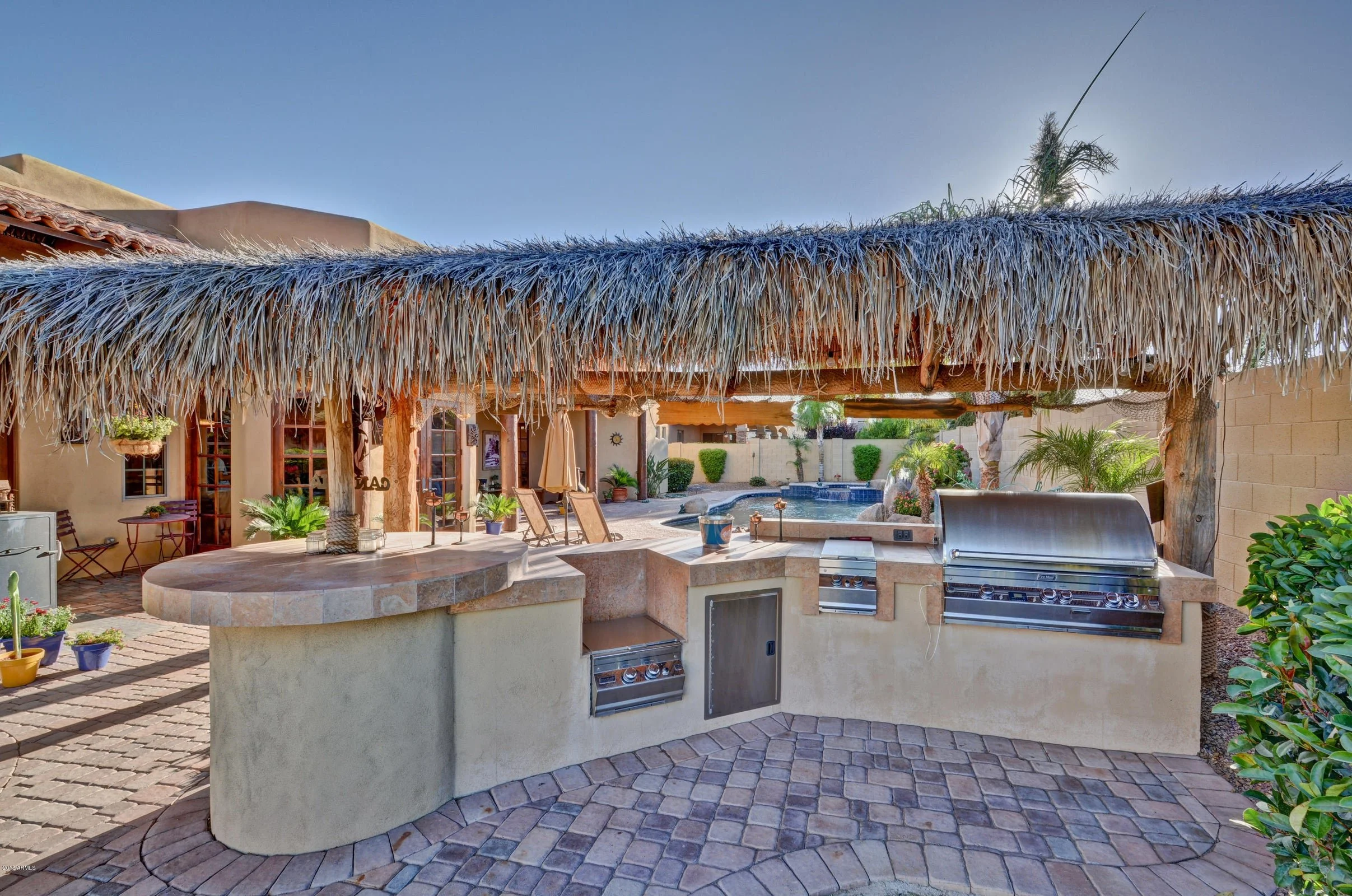 Outdoor kitchen with built-in grills under a thatched roof, with a swimming pool, lounge chairs, plants, and patio furniture in the background.