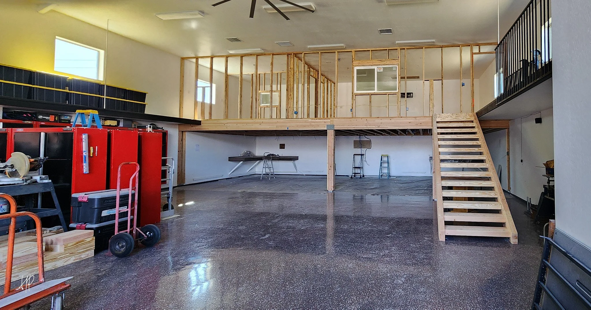 Interior of a building under construction with an upper level and wooden staircase. Construction tools and supplies are scattered on the floor and a work table.