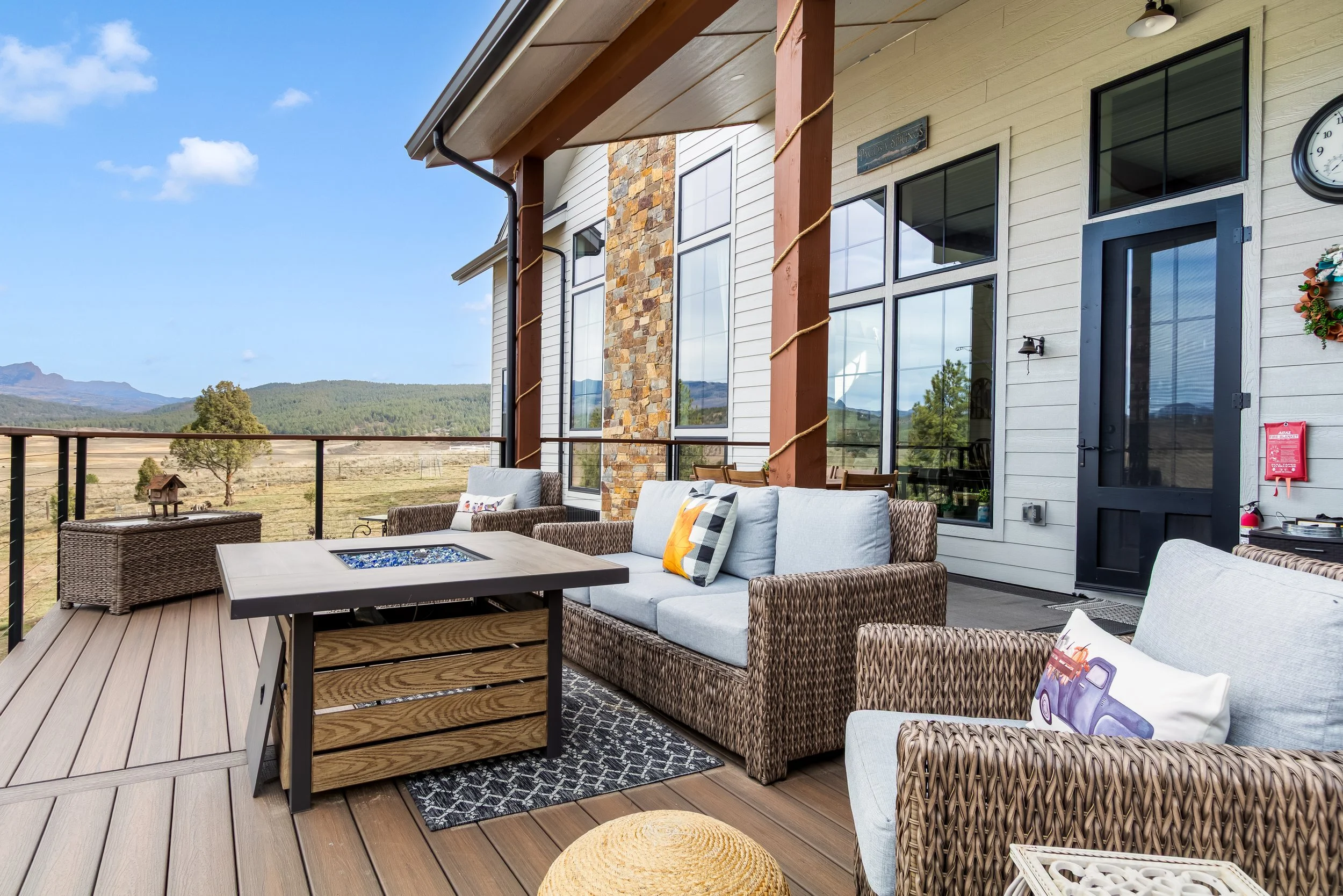 Outdoor balcony with wicker furniture, cushions, a fire pit table, and scenic mountain view.