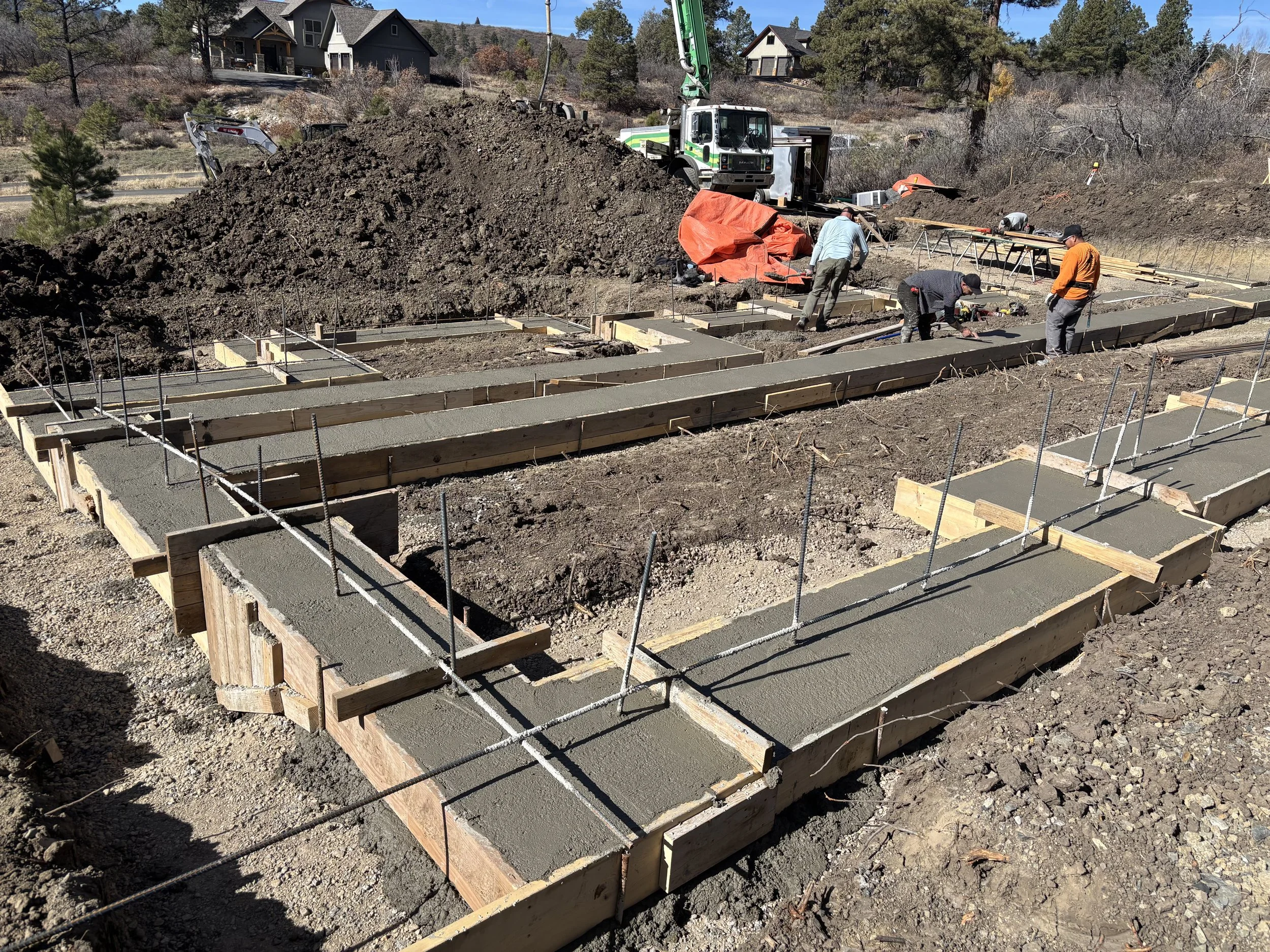 Construction site with concrete foundation forms being set for a building, workers are working on the foundation, with a truck and construction equipment in the background.