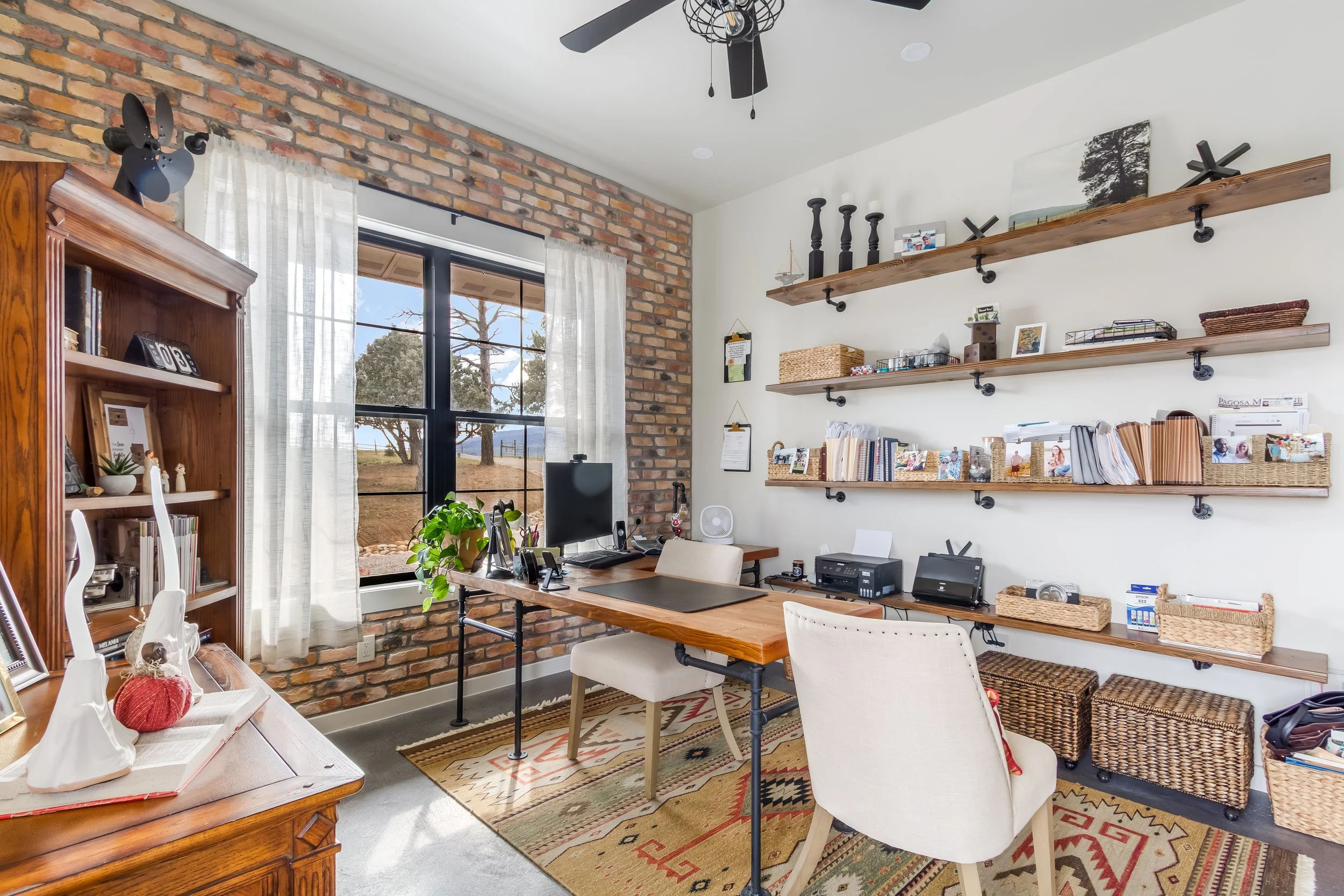 Home office with brick wall, large window with white curtains, wooden desks, white chairs, wall-mounted shelves with decor and books, wicker baskets, patterned rug, ceiling fan, and outdoor view with trees.