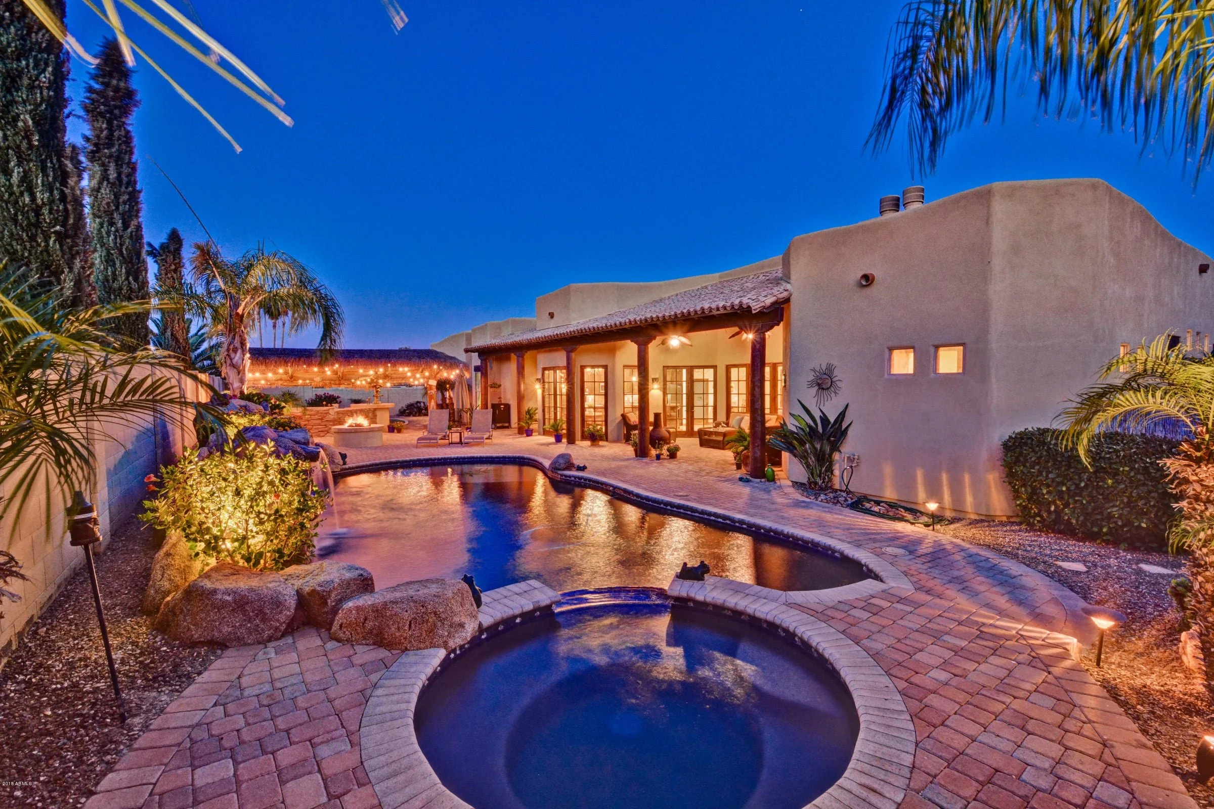 A backyard with a swimming pool and hot tub under a clear evening sky, surrounded by palm trees and outdoor furniture, with a house illuminated from inside.