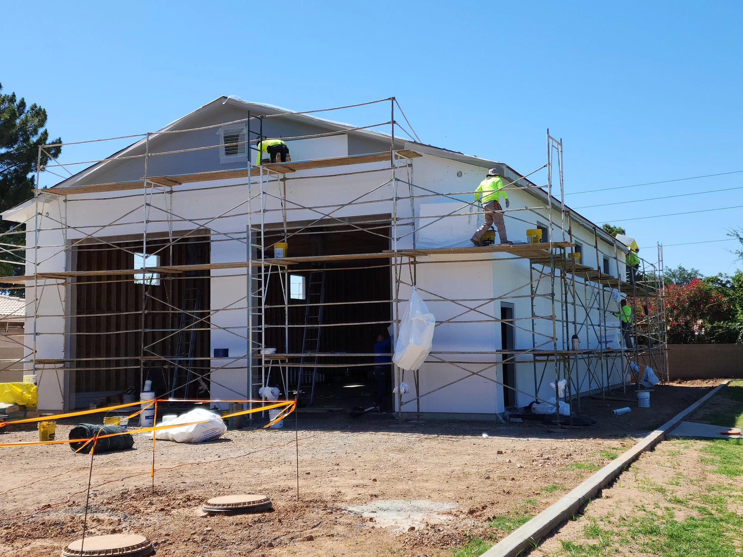 Construction workers on scaffolding working on a two-story house with white exterior walls and a gable roof, under a clear blue sky.