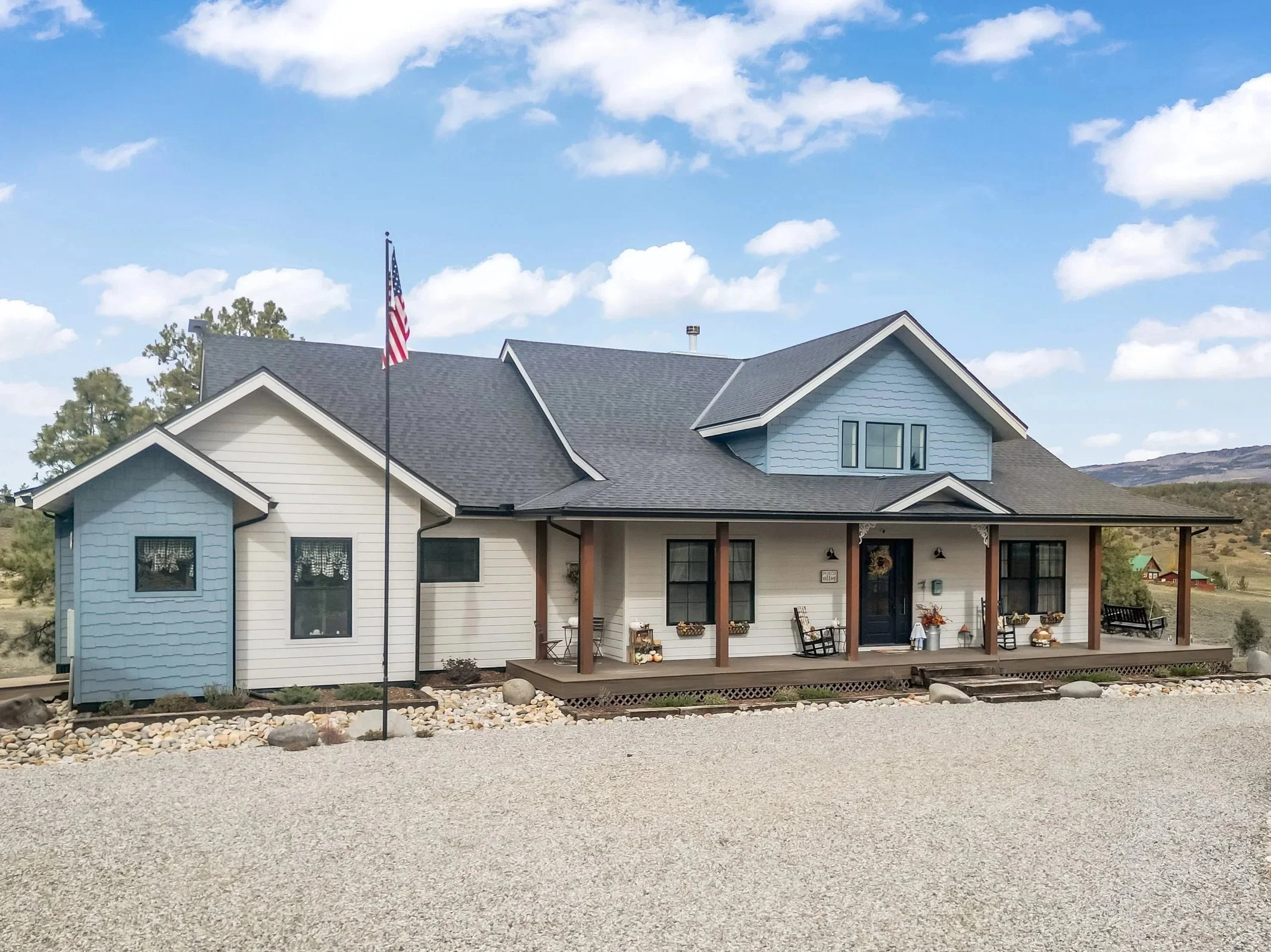 A custom house with a front porch, dark door, blue and white exterior, American flag, surrounded by gravel and rocks, in a rural setting with hills in the background and a partly cloudy sky.