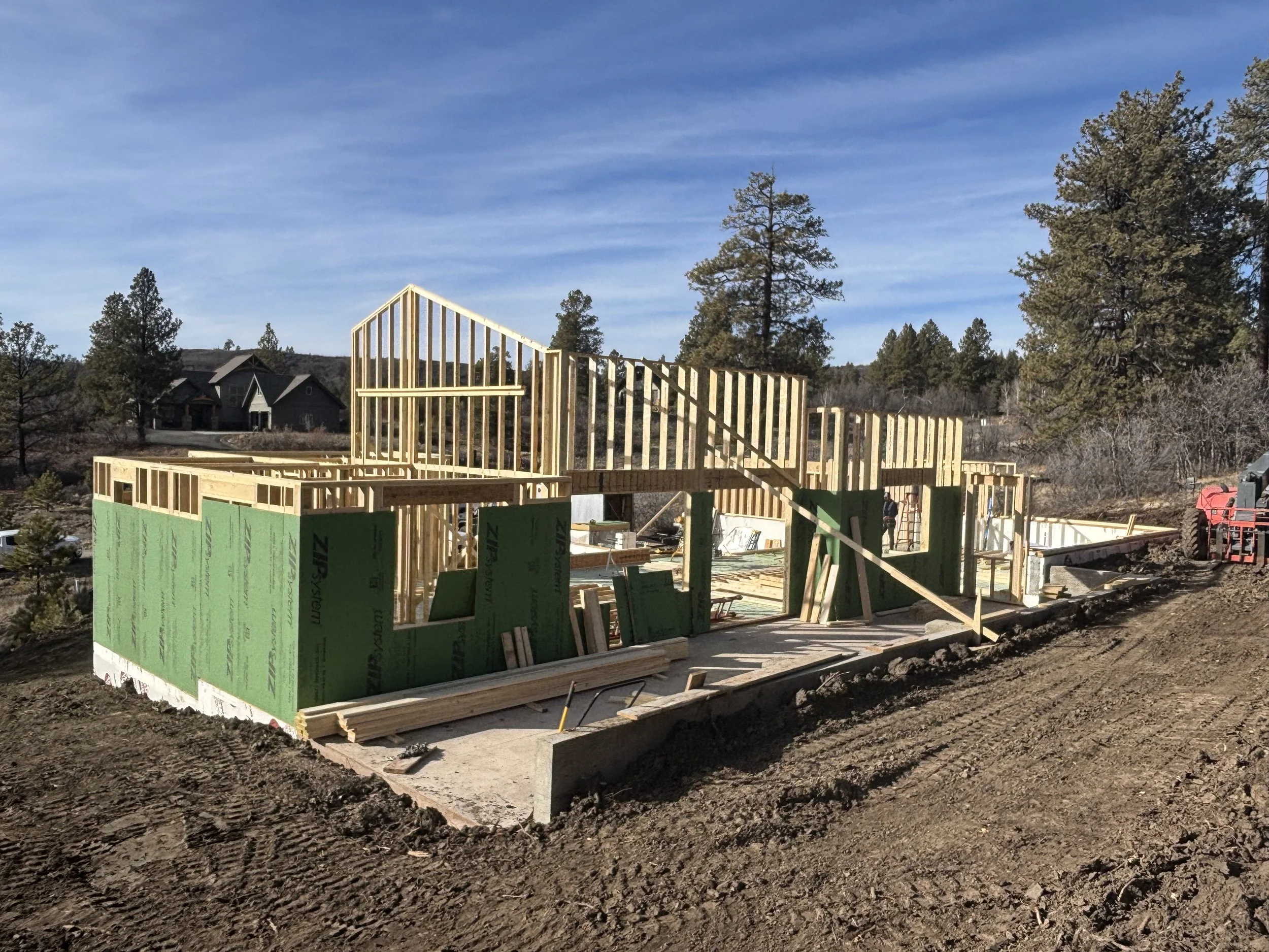 Construction site of a house with wooden framing and green insulation panels, surrounded by dirt and trees, under a clear blue sky.