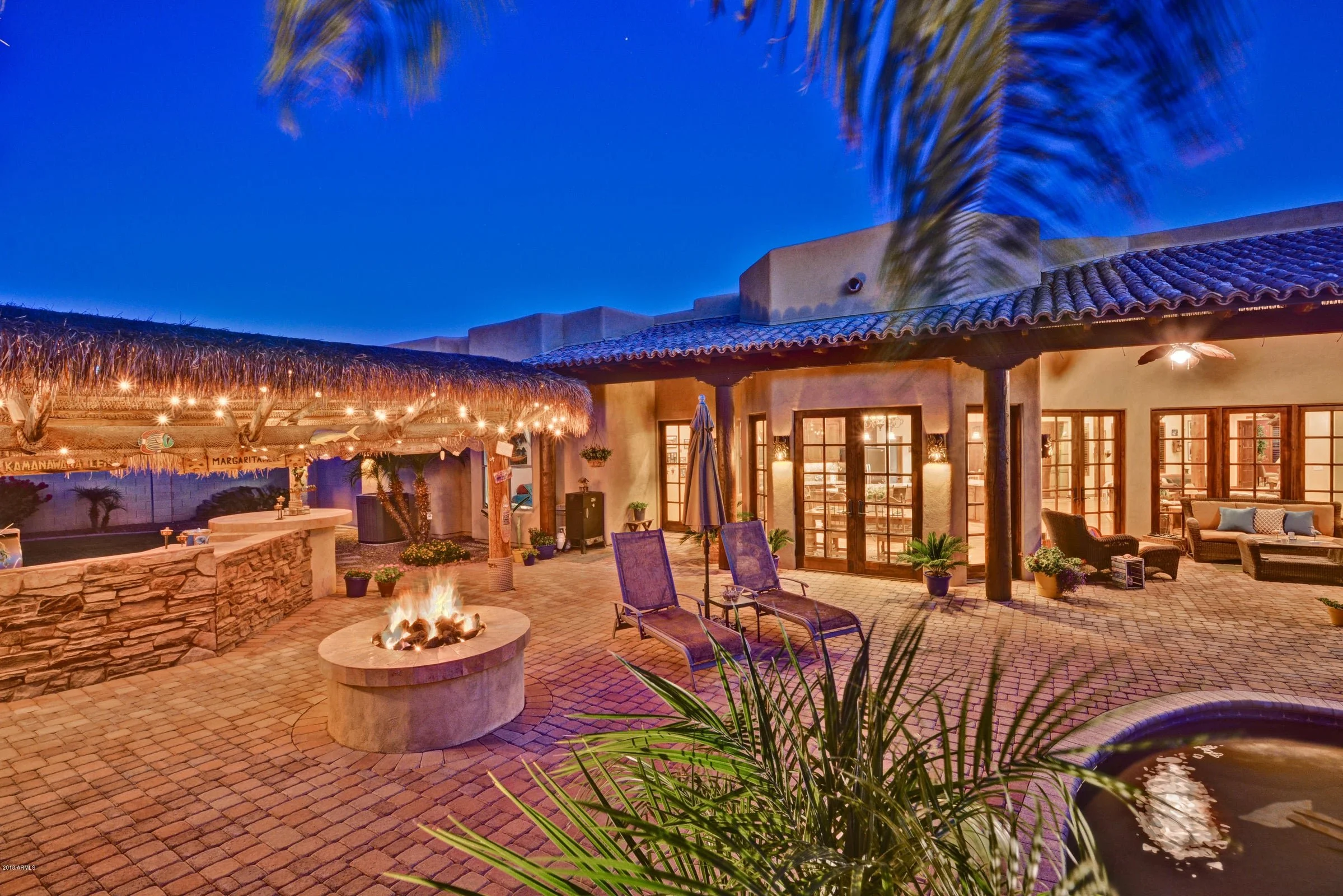 An outdoor patio at dusk with a fire pit, lounge chairs, and cozy seating area under a thatched roof, surrounded by plants and string lights.