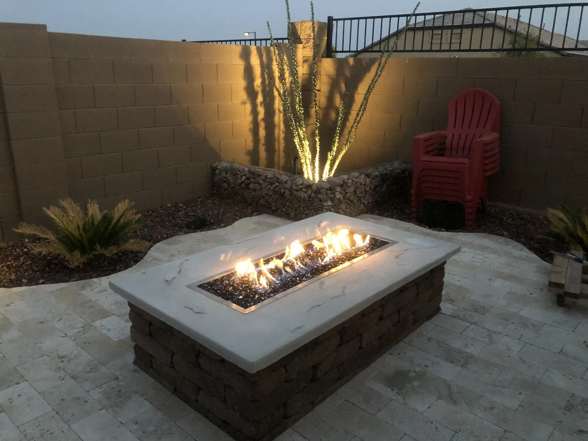A backyard patio at dusk with a rectangular fire pit, a small palm plant, stacked red chairs, a beige brick wall, and a lit cactus with shadows cast on the wall.