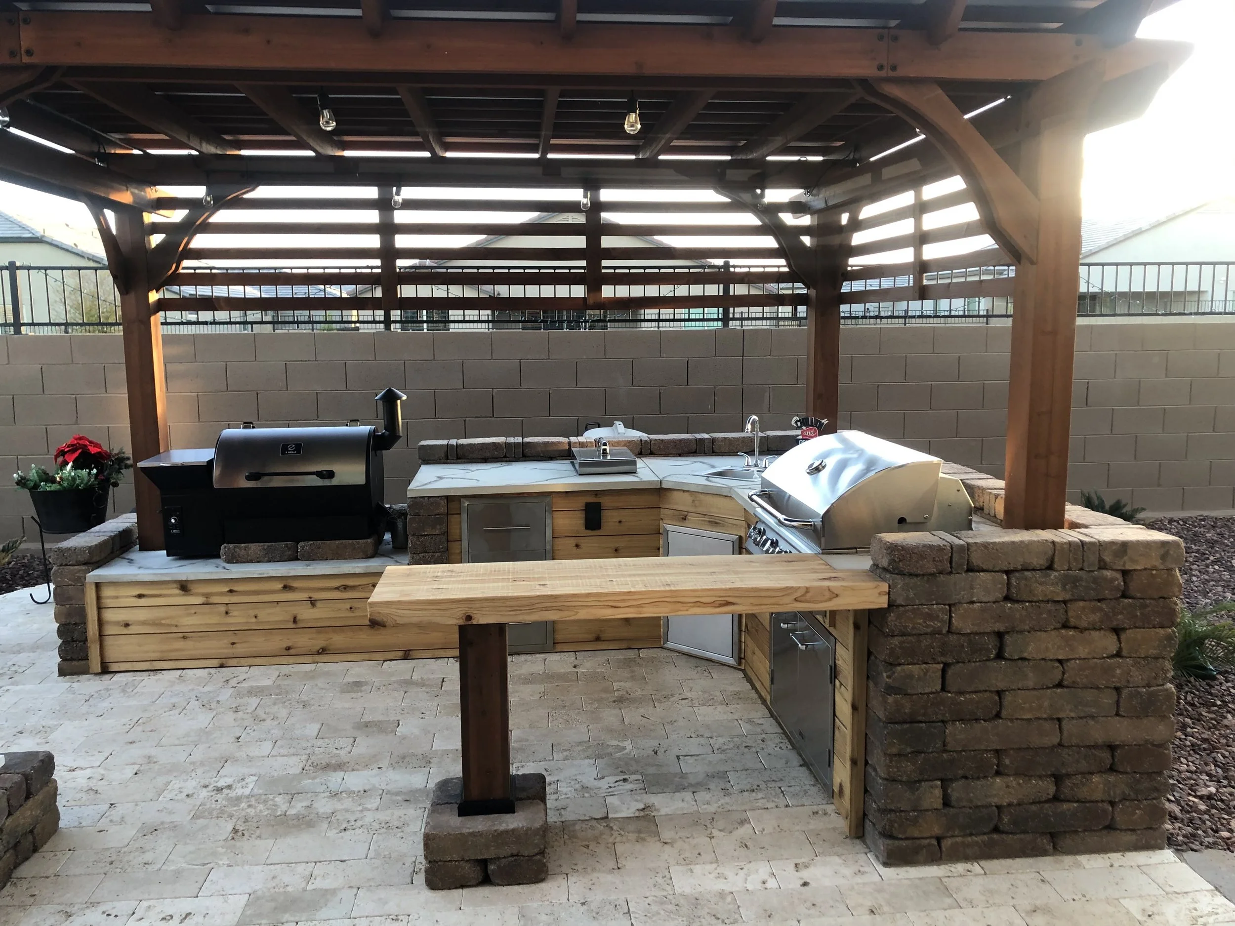 Outdoor kitchen area with a brick and wood structure, featuring a grill, a smoker, a small sink, and storage cabinets, under a wooden pergola.