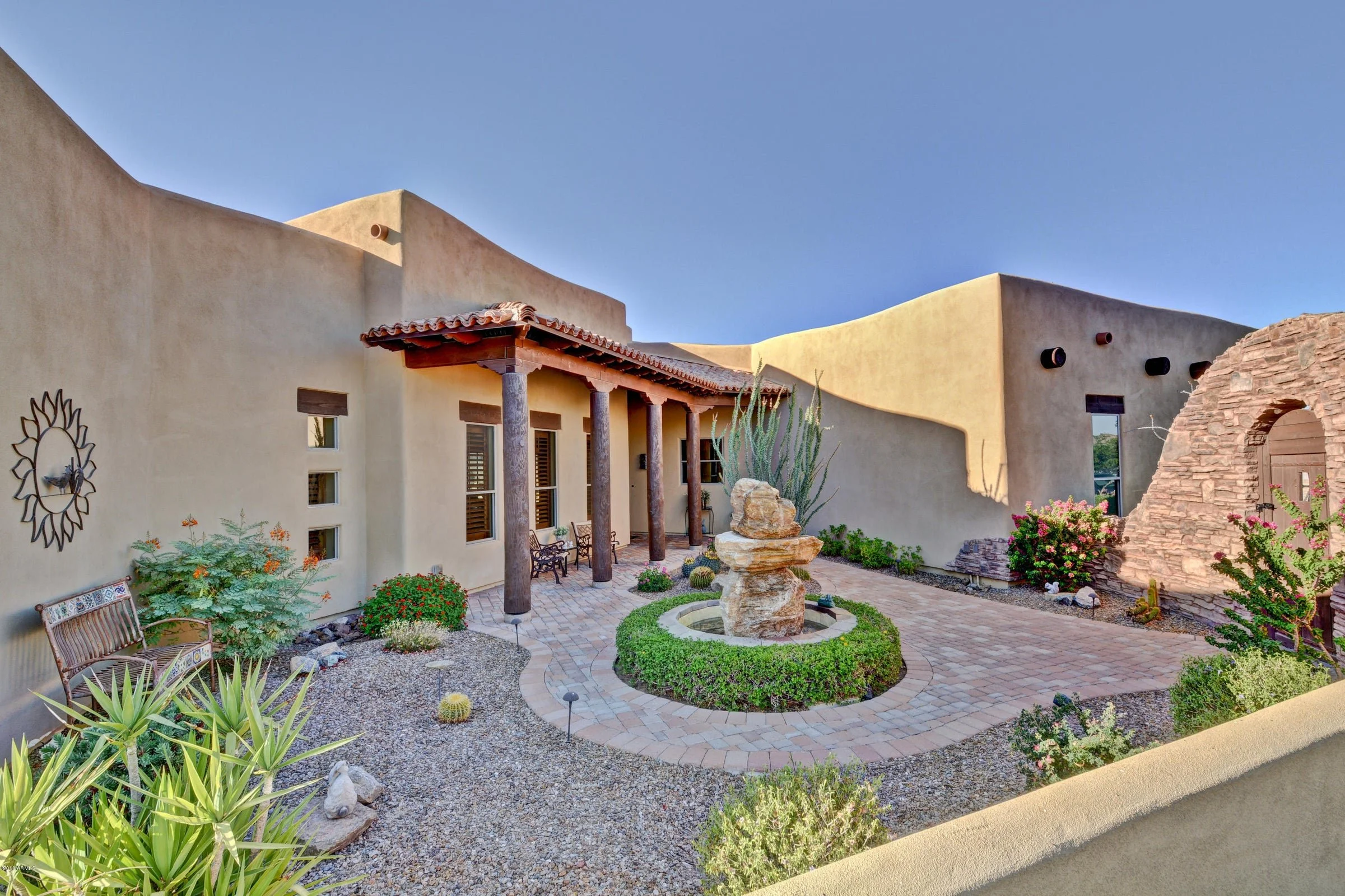 Mexican-style courtyard with beige stucco walls, a brick walkway, desert plants, and a central rock fountain with a cactus plant