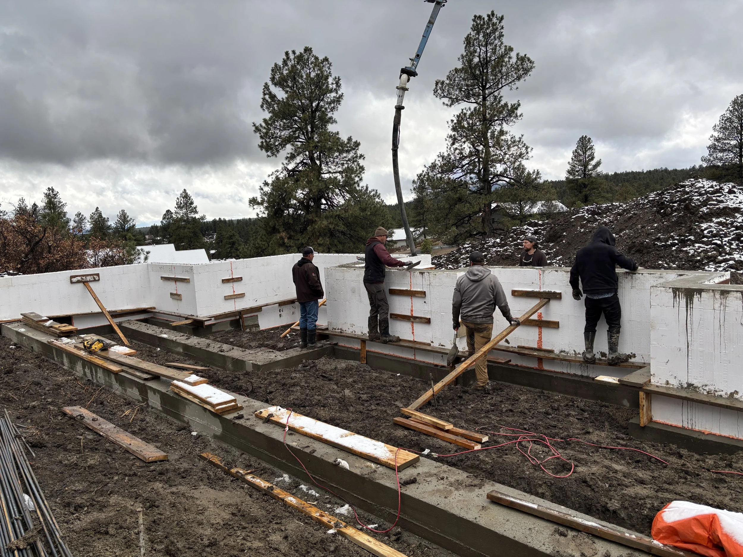 Construction workers build a concrete wall on a muddy site with trees and cloudy sky in background.