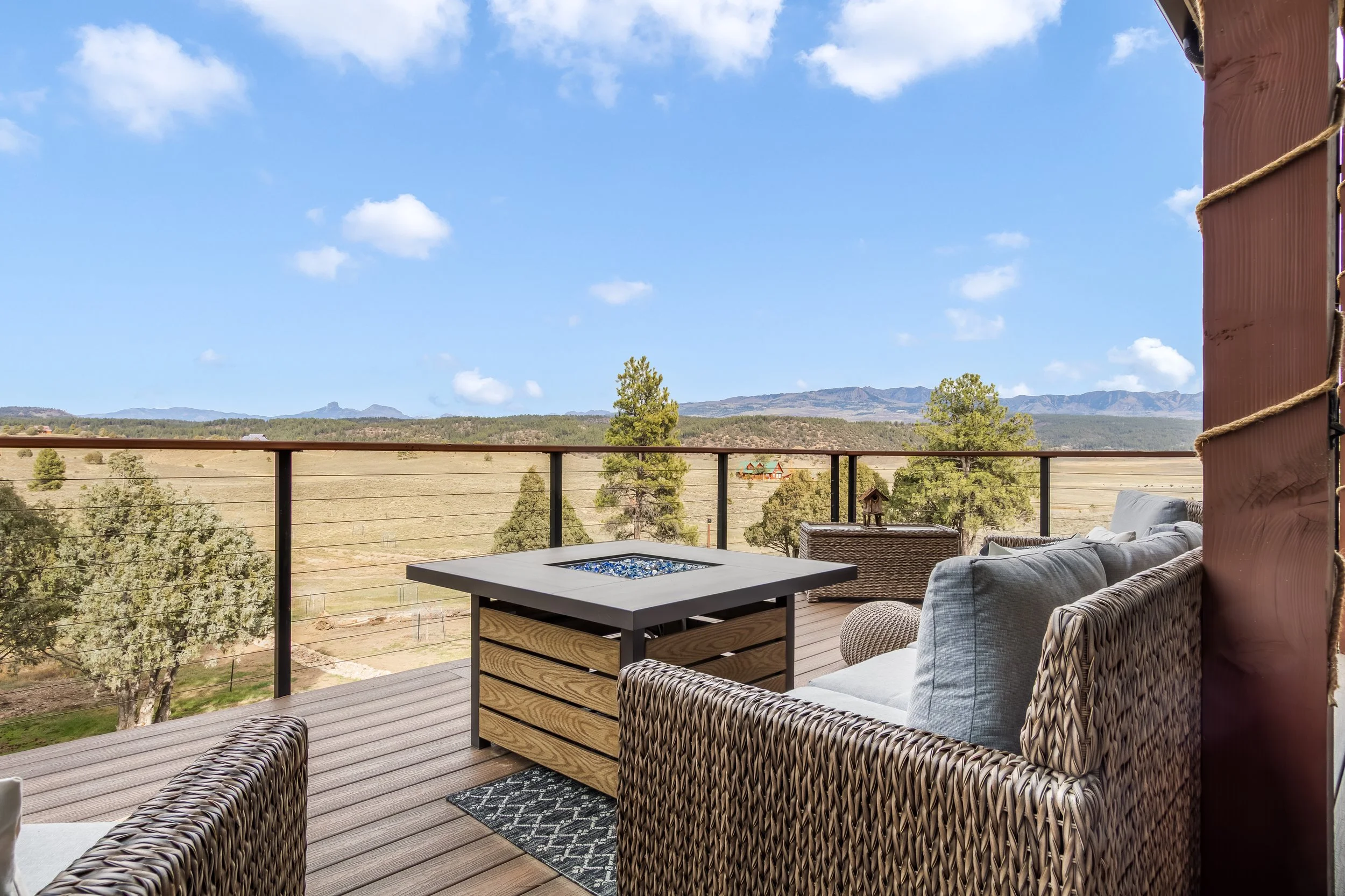 Patio with wicker furniture and a view of trees, hills, and mountains under a blue sky with scattered clouds.