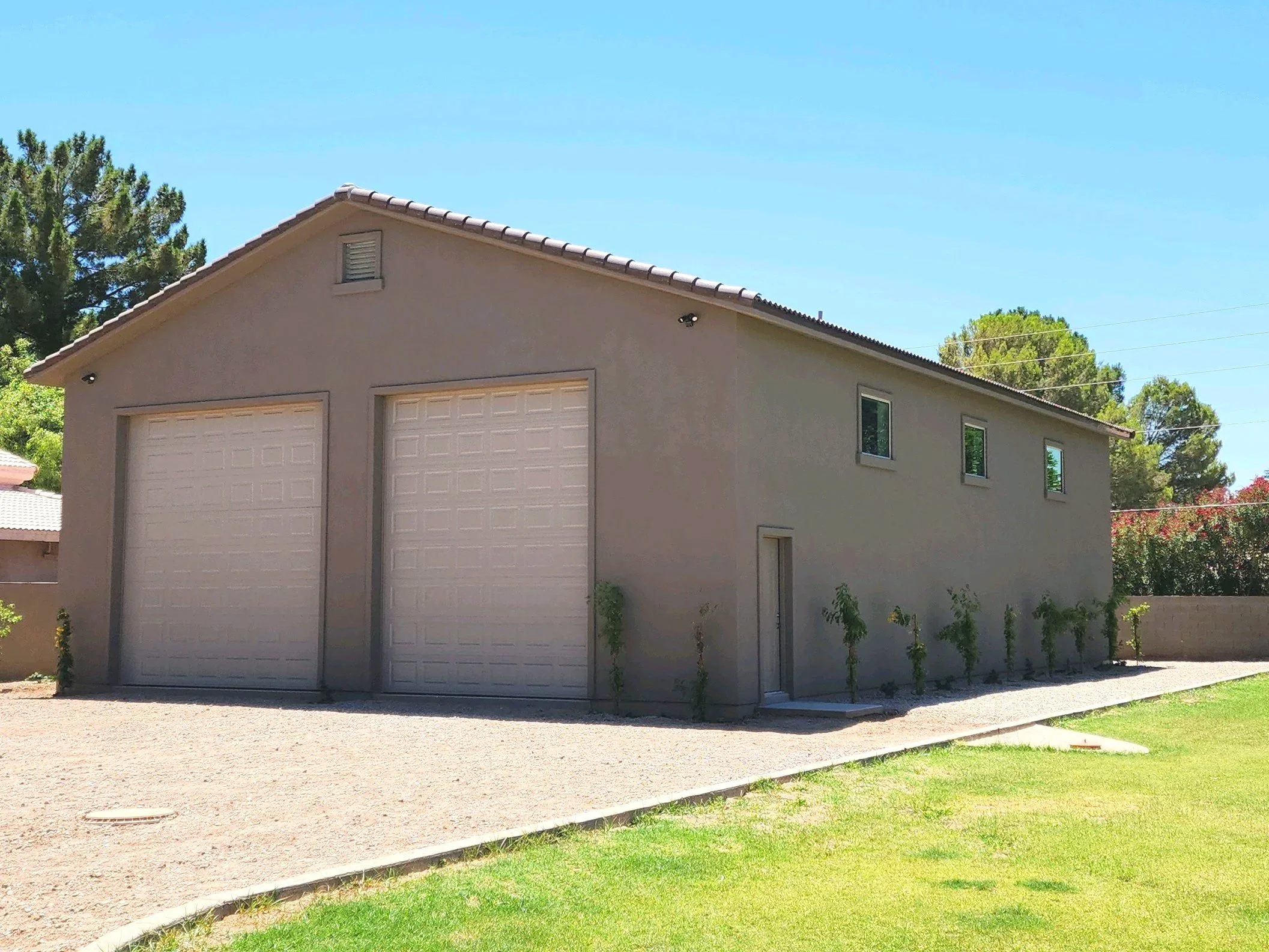A two-story beige garage building with two white garage doors and a side door, surrounded by a gravel driveway, small plants, and a grassy lawn, under a clear blue sky.