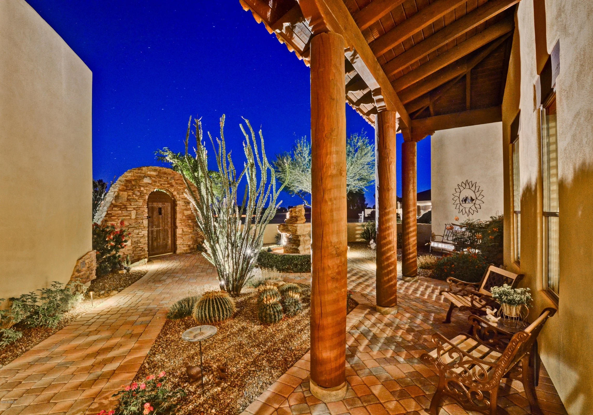 Nighttime view of a southwestern-style porch with brick flooring, wooden columns, a cactus, and desert plants illuminated by landscape lighting. The sky is dark blue with stars visible.