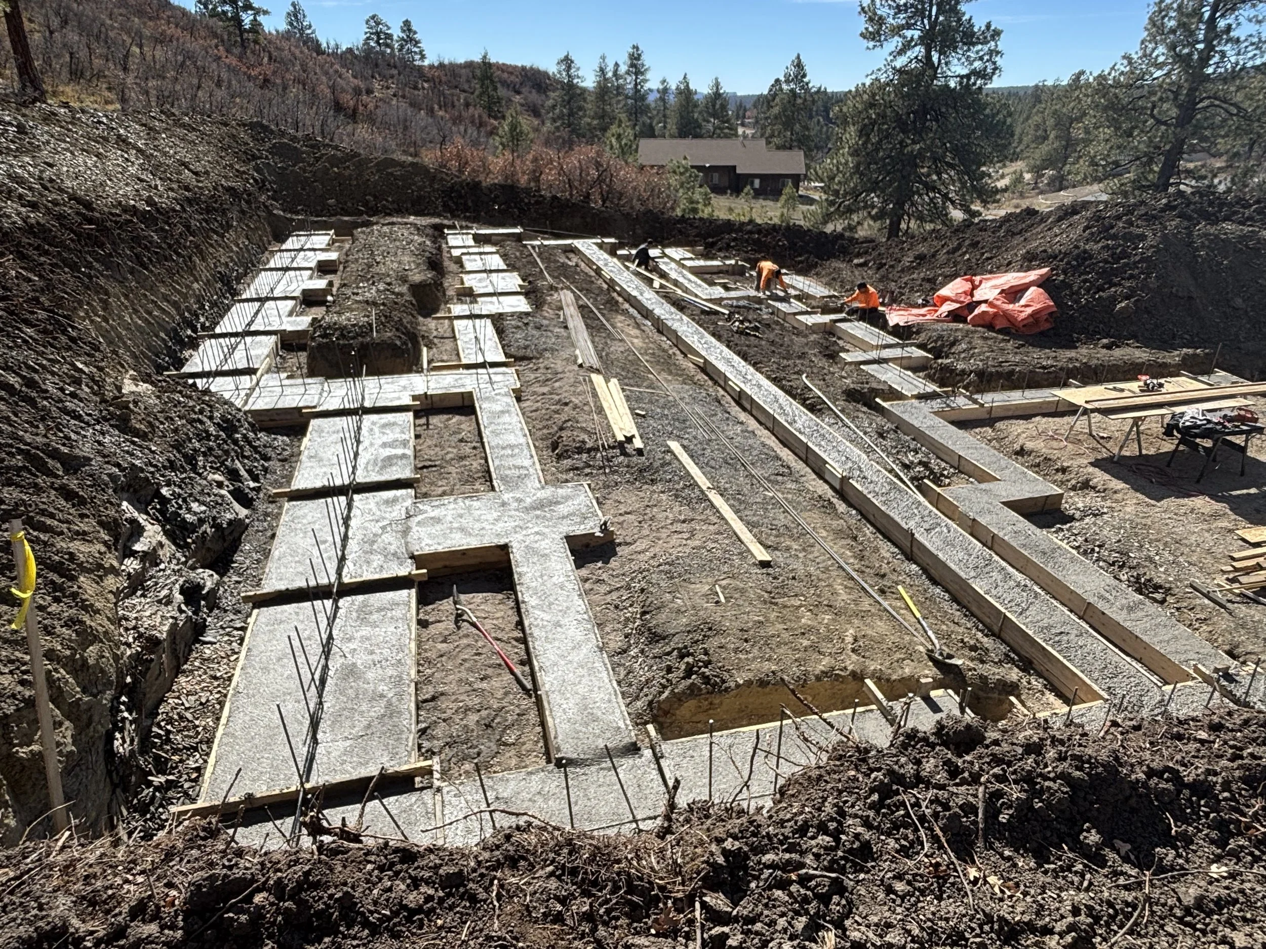 Construction site with workers pouring concrete foundation on hilly terrain, with trees and a house in the background.