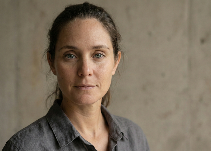 A woman with light skin and brown hair tied back, wearing a gray button-up shirt, standing against a plain concrete wall.