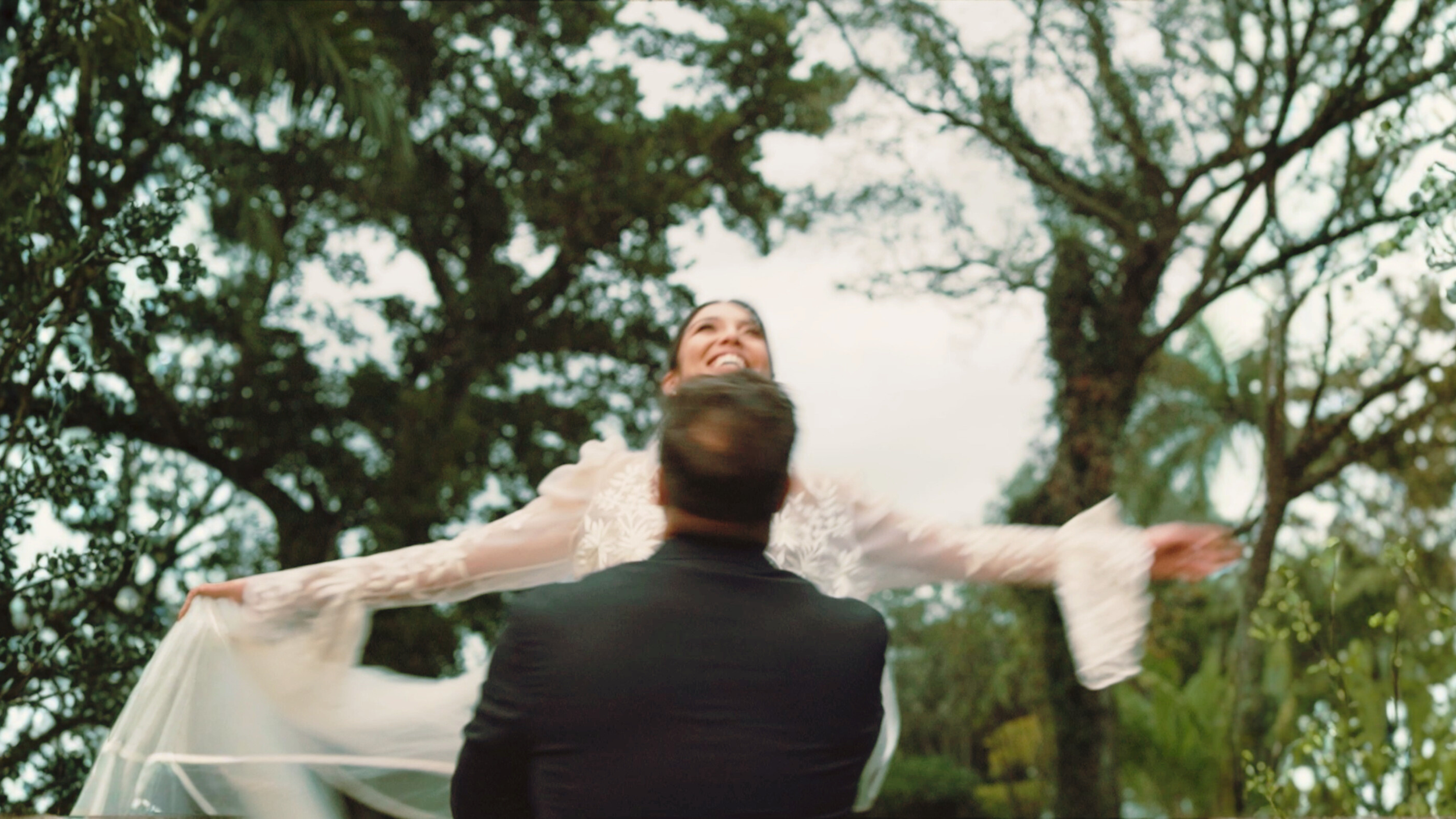 A woman in a wedding dress being lifted by a man in a suit outdoors among trees, with her arms outstretched and smiling.