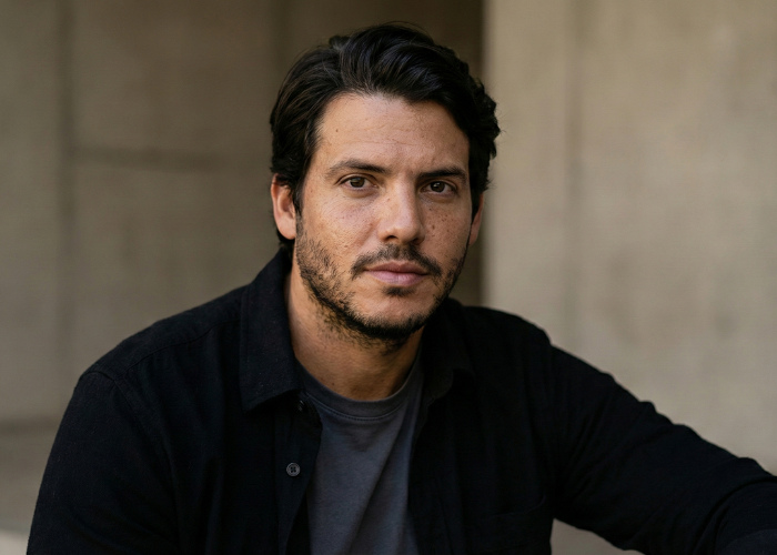 A man with dark hair and facial stubble looking at the camera outdoors, with a neutral expression and a dark shirt.