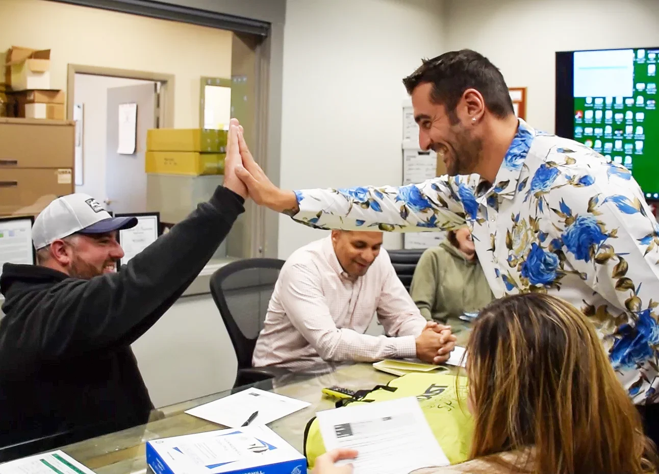Two men give each other a high-five and smile in a meeting room with four other people.