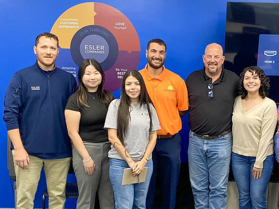 Six people standing in front of a blue wall with a circular chart and text. The group includes three men and three women, smiling and posing for the photo.