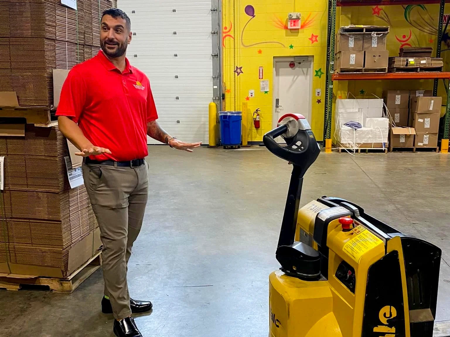 A man in a red polo shirt, beige pants, and black shoes stands with his hands raised in front of a yellow pallet jack in a warehouse. The warehouse has cardboard boxes on pallets, a yellow wall with decorations, and shelves with boxes in the background.
