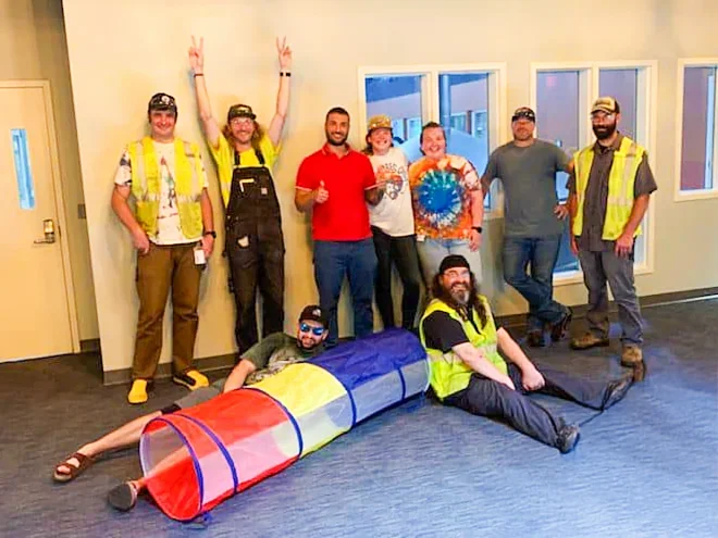 Group of people posing indoors with some wearing safety vests, one person lying on the floor in a fabric tunnel, and others standing behind them, some making peace signs, in a room with large windows.