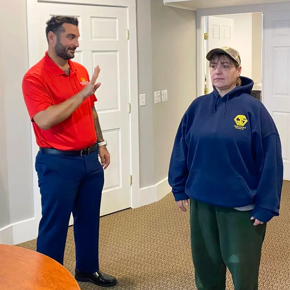A man in a red polo shirt and a woman in a blue hoodie standing in a room with beige walls and white doors, with the man raising his hand in a greeting gesture.