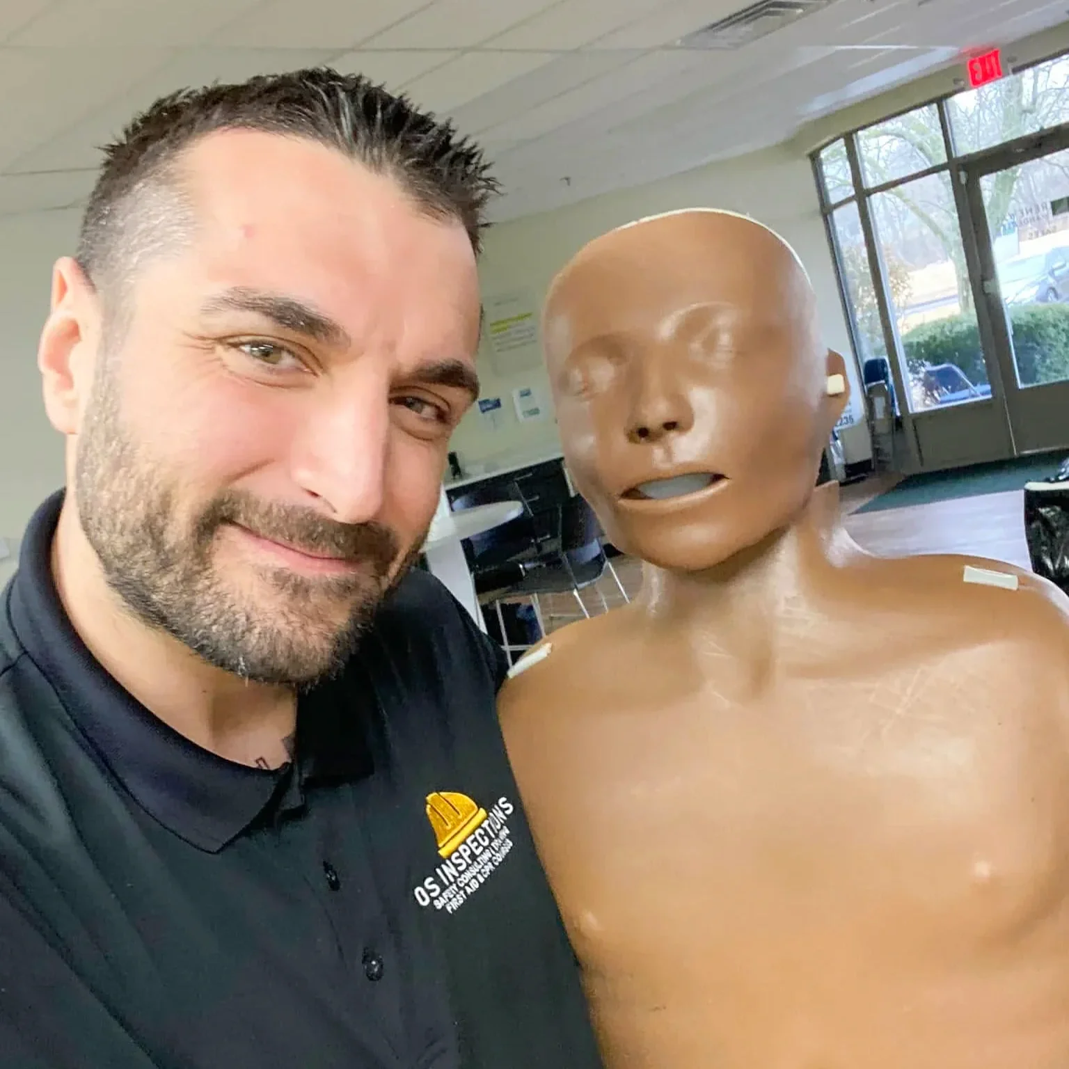 A man taking a selfie with a life-size medical training mannequin in an indoor setting with large windows.