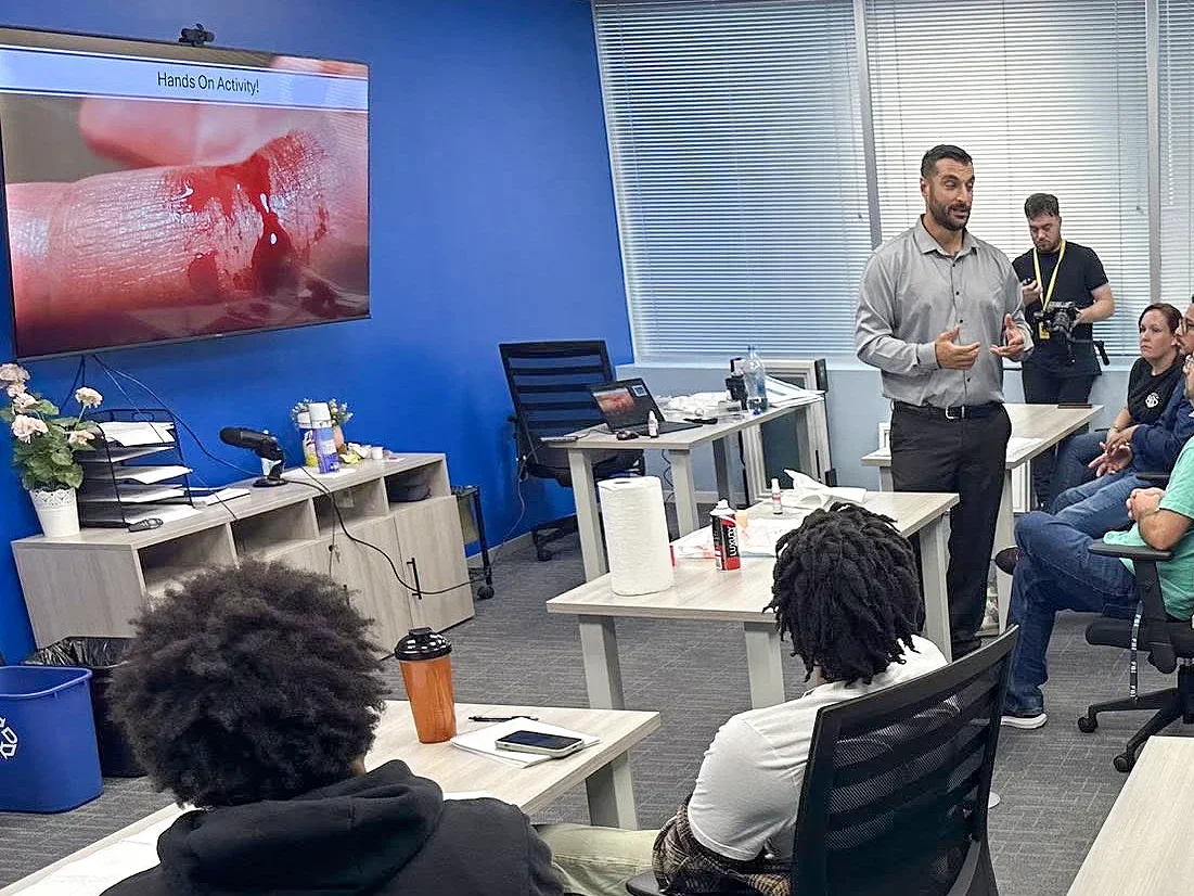 A man giving a presentation with a slide showing a close-up of a finger with a cut and blood. The room has several people seated, and a large screen displays the presentation. There is equipment and supplies on the tables, including disinfectant bottles and paper towels.