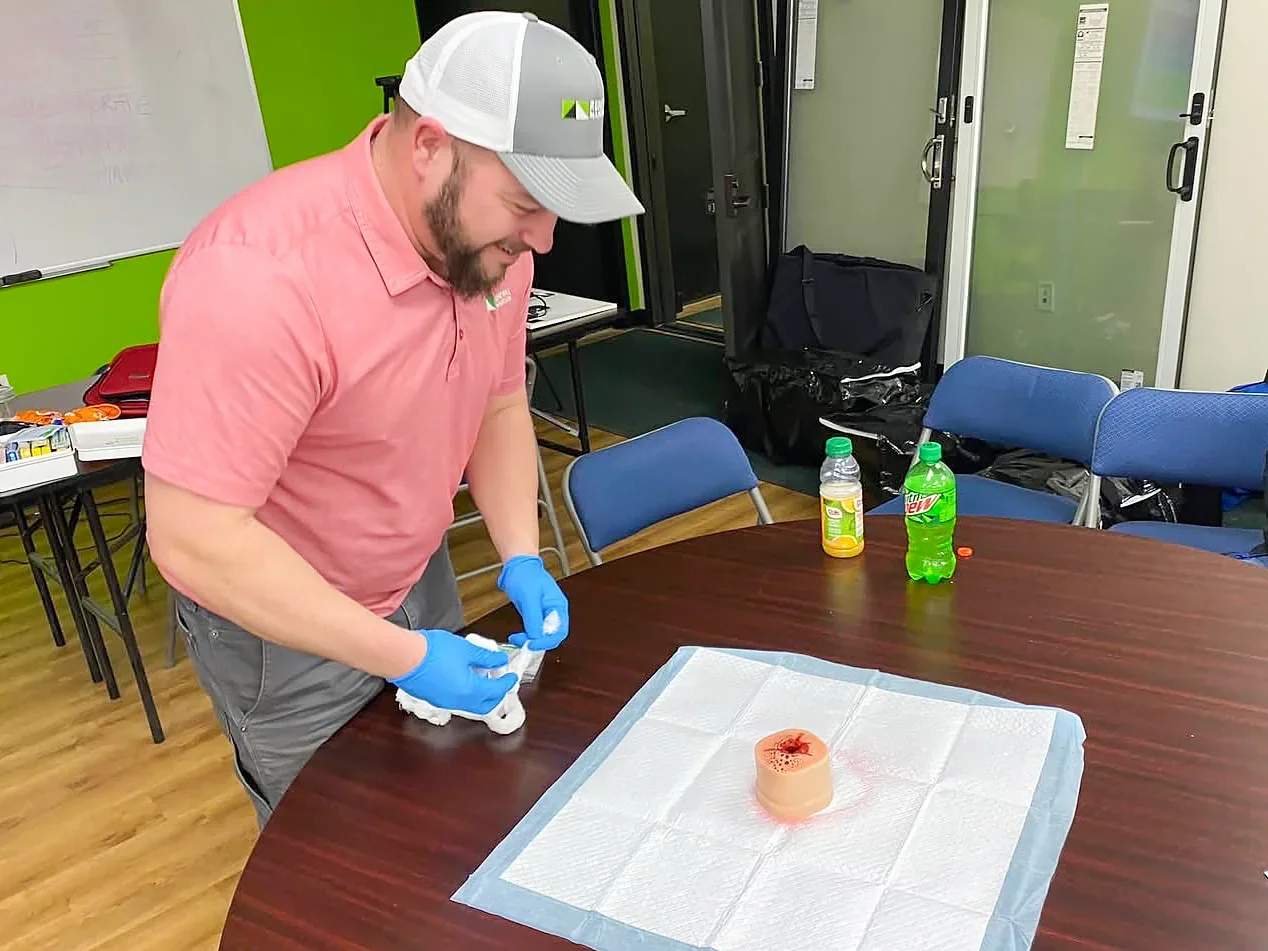 A man in a pink shirt and gray cap with a beard, wearing blue gloves, is working with medical supplies at a table with a pad and a model of a human organ. The room has green and white walls, folding chairs, bottles of tea and soda on the table, and various equipment and bags in the background.