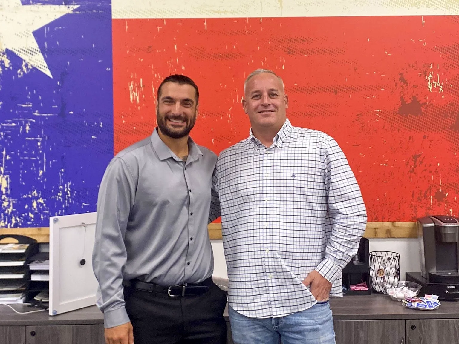 Two men standing indoors in front of a large American flag mural, smiling at the camera.