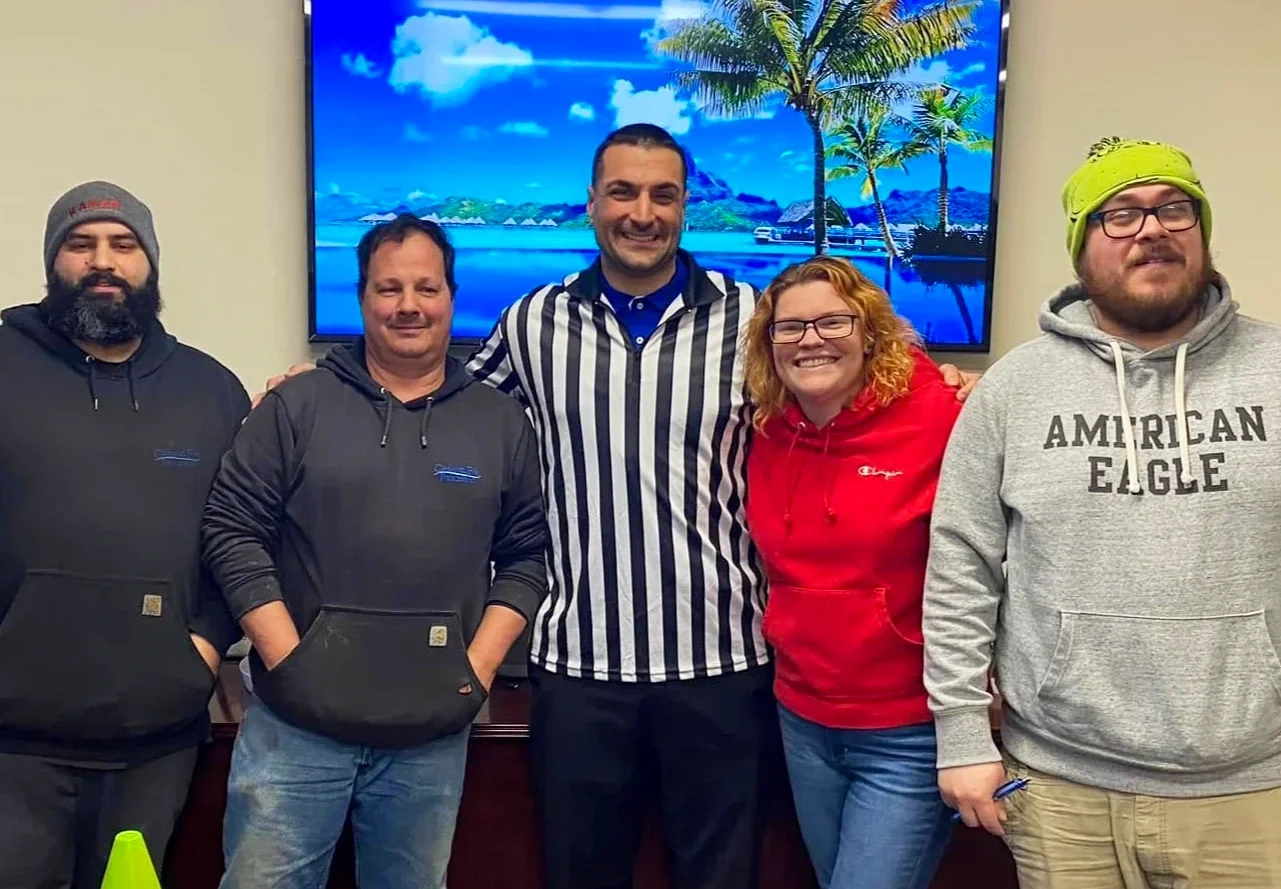 Group of five people standing indoors, smiling at the camera, with a tropical beach scene on a television screen behind them.